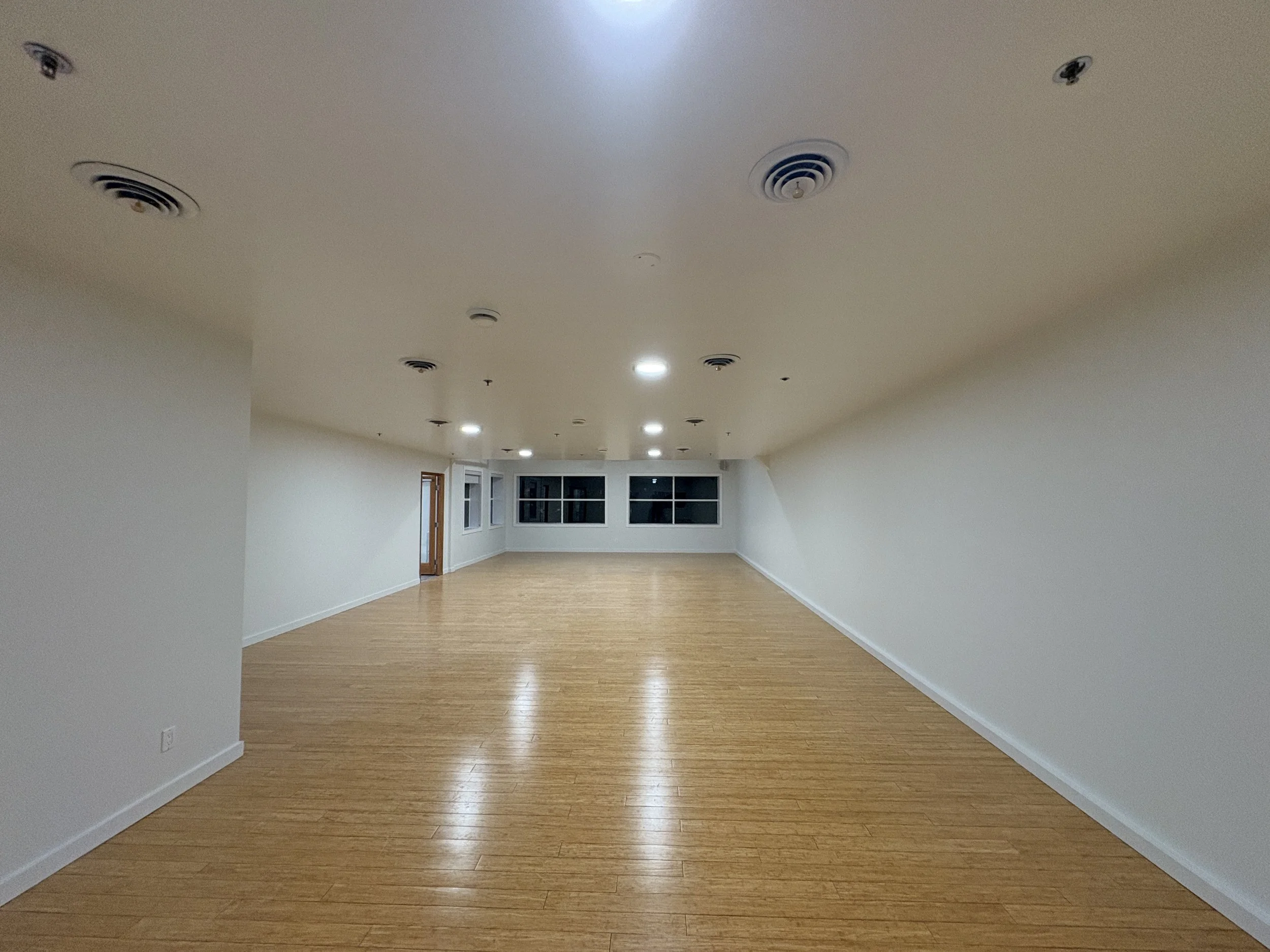 Empty room with wooden flooring, white walls, recessed lighting, and a window at the far end.