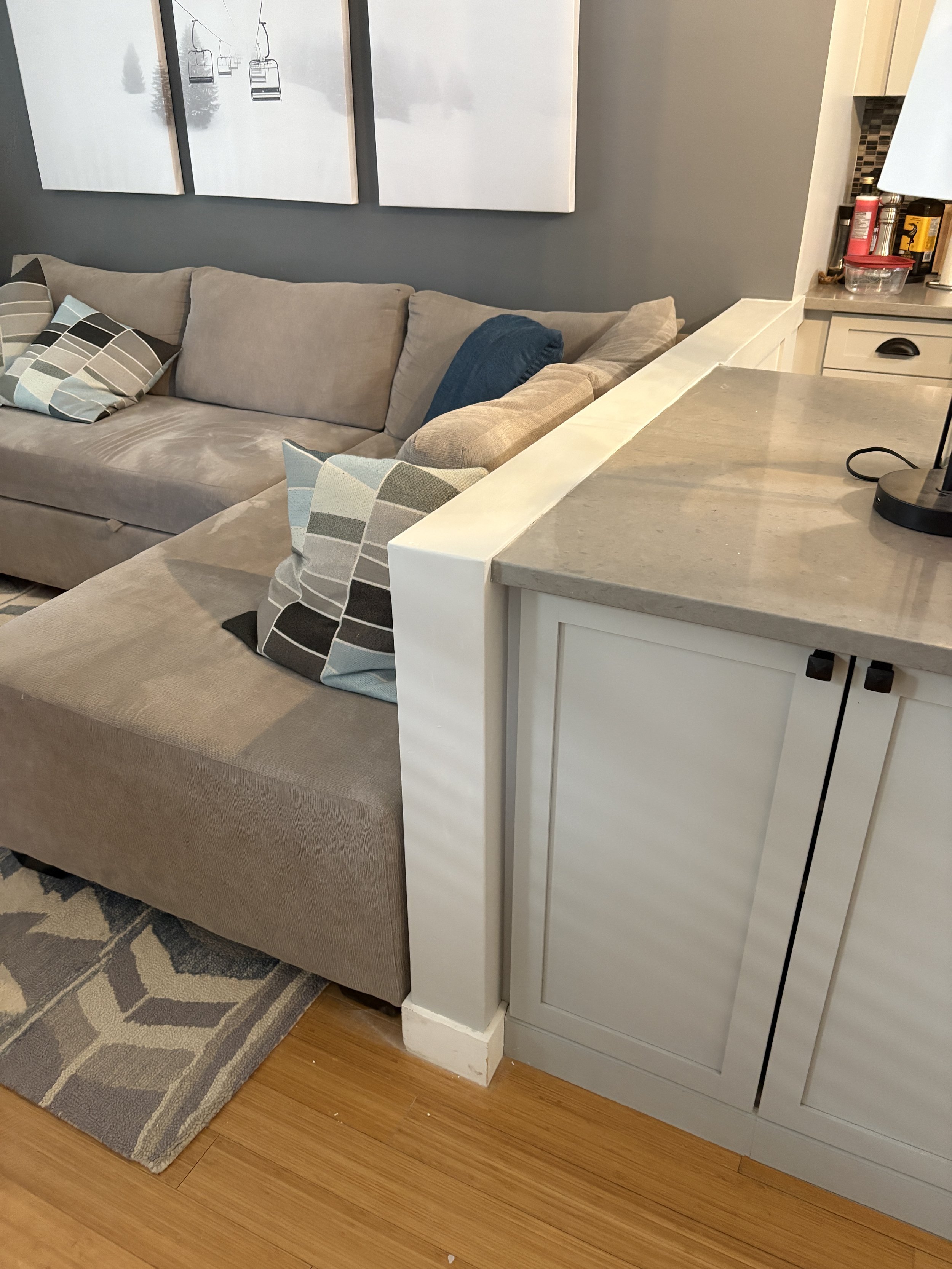 Living room with gray sectional sofa, geometric patterned pillows, wall art of a snowy landscape, light wood flooring, and a white cabinet beside a countertop.