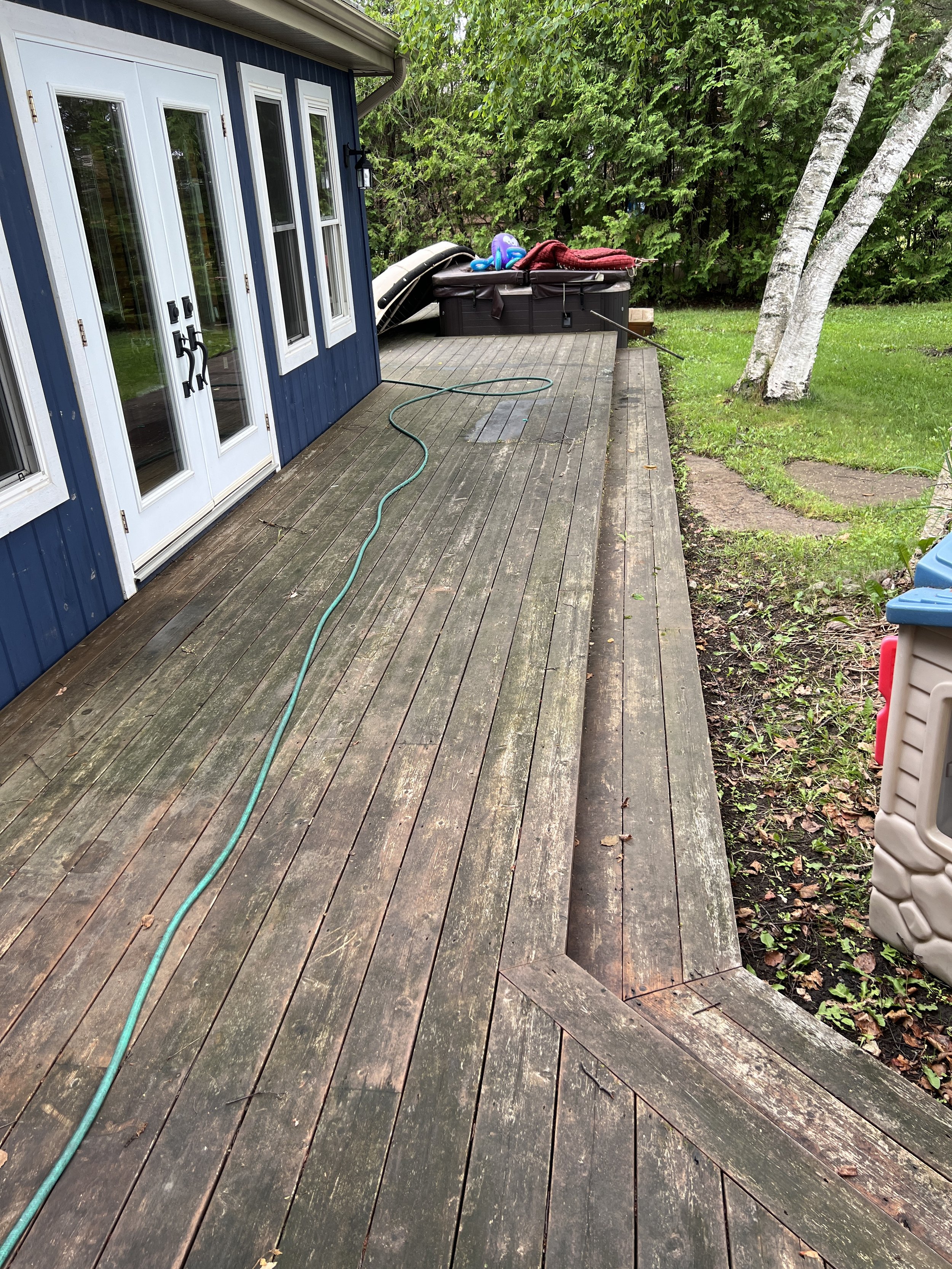Wooden deck with hose and patio furniture, surrounded by greenery and trees, beside a blue house with white doors and windows.