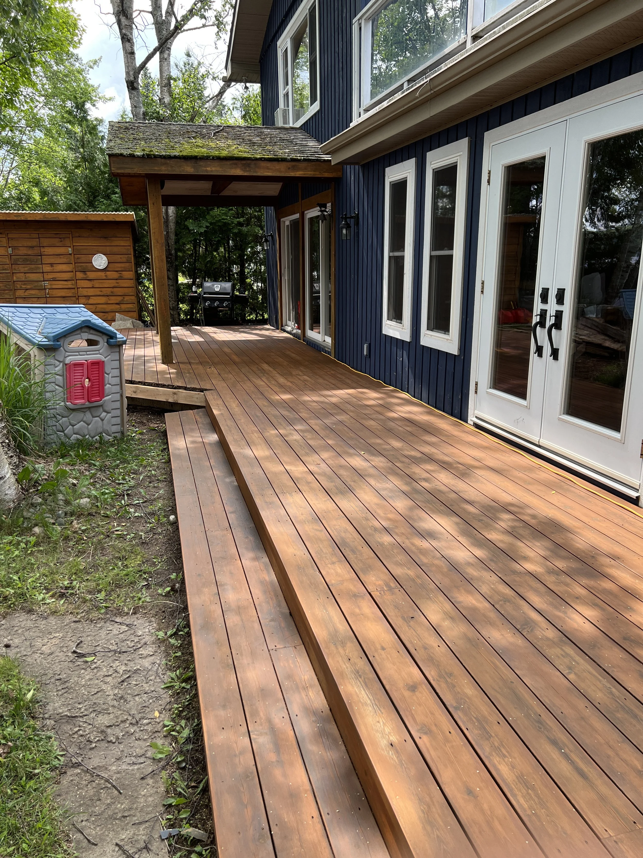 Photo of a backyard with a wooden deck attached to a blue house with white-framed glass doors and windows. There is a small covered area with a mossy roof, a barbecue grill, and a children's plastic playhouse nearby. The yard is surrounded by trees a