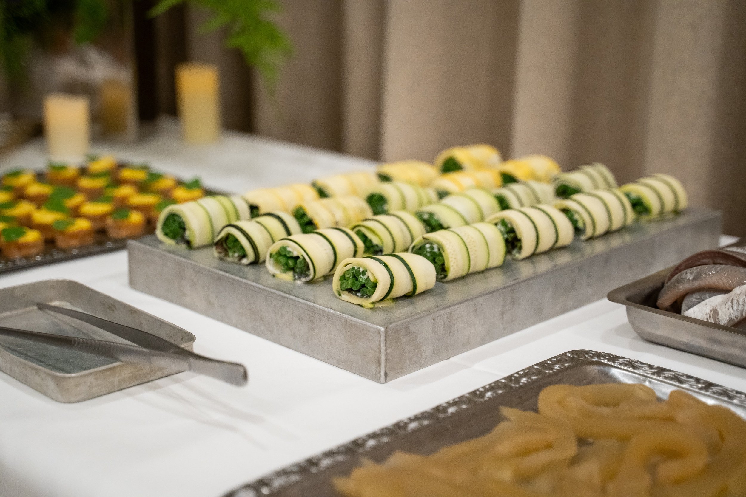 Tube-shaped cucumber roll appetizers with green filling on a metal tray, with other assorted appetizers in the background.