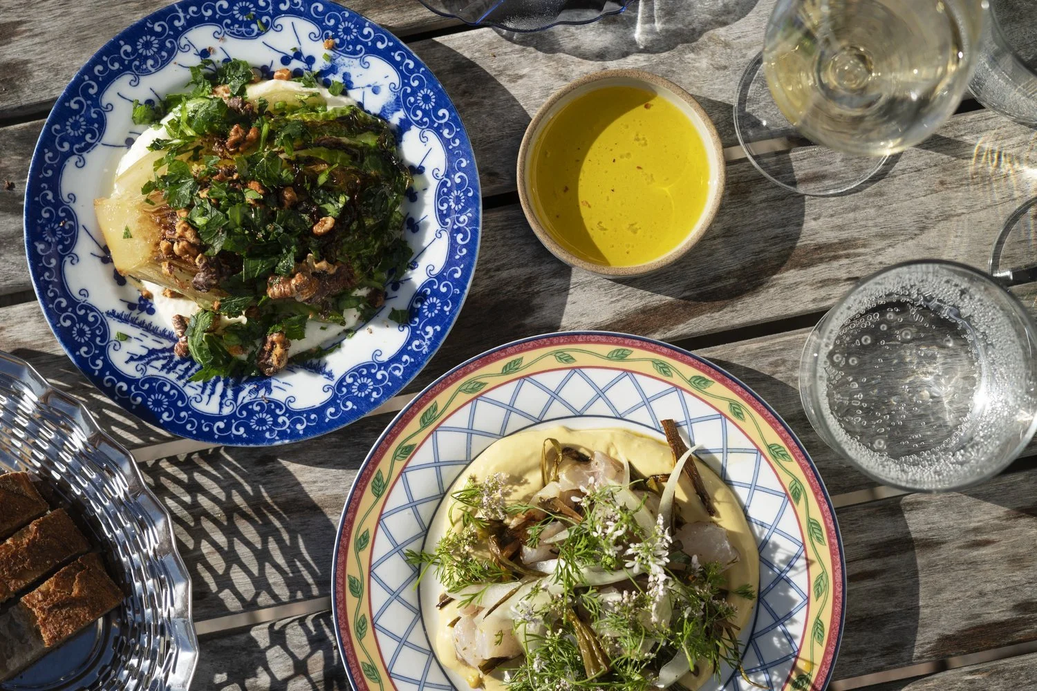 A variety of dishes and drinks on a wooden table, including a blue-rimmed plate with baked potato topped with chopped herbs and possibly meat, a colorful plate with a seafood dish garnished with herbs and edible flowers, a bowl of yellow sauce or sou