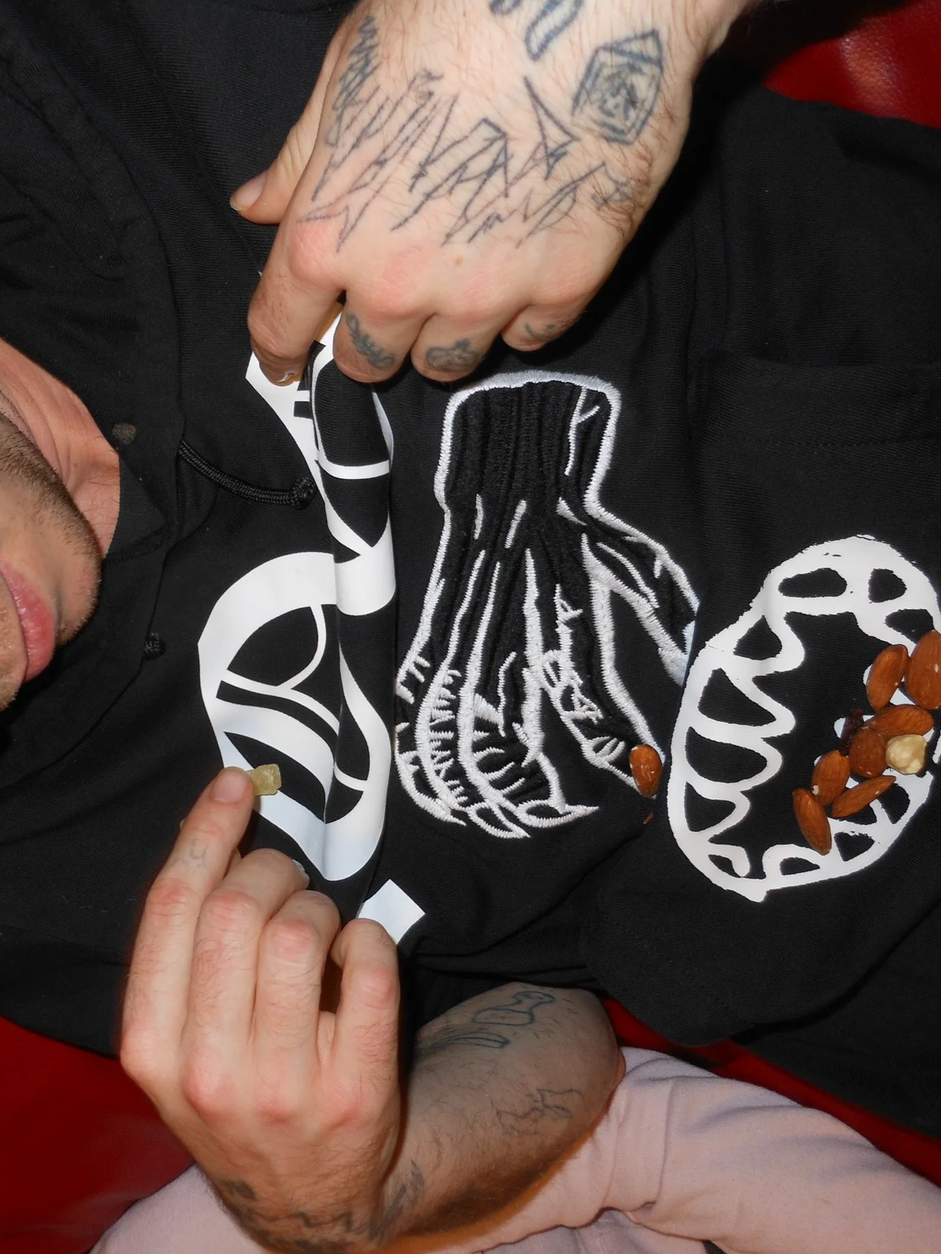 Close-up of a person wearing a black hoodie with white embroidered designs, lying down with a few almonds in their hand and some almonds scattered on their hoodie. The person has tattoos on their hands and arms.