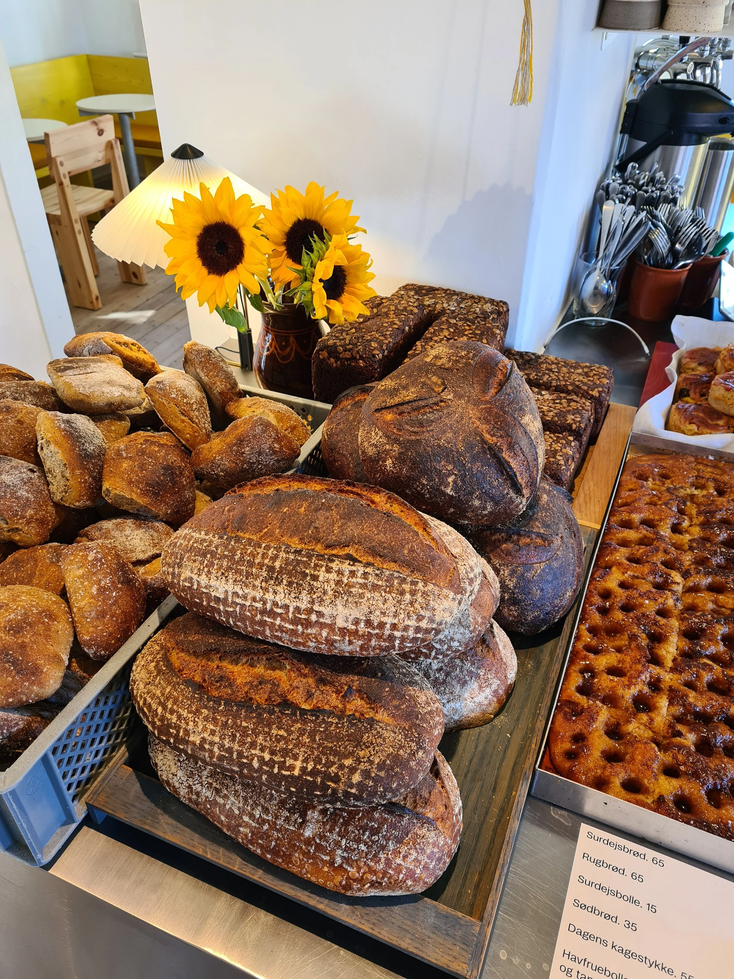 Assorted freshly baked bread loaves and rolls displayed on a counter in a bakery, with a small sunflower bouquet and a desk lamp in the background.