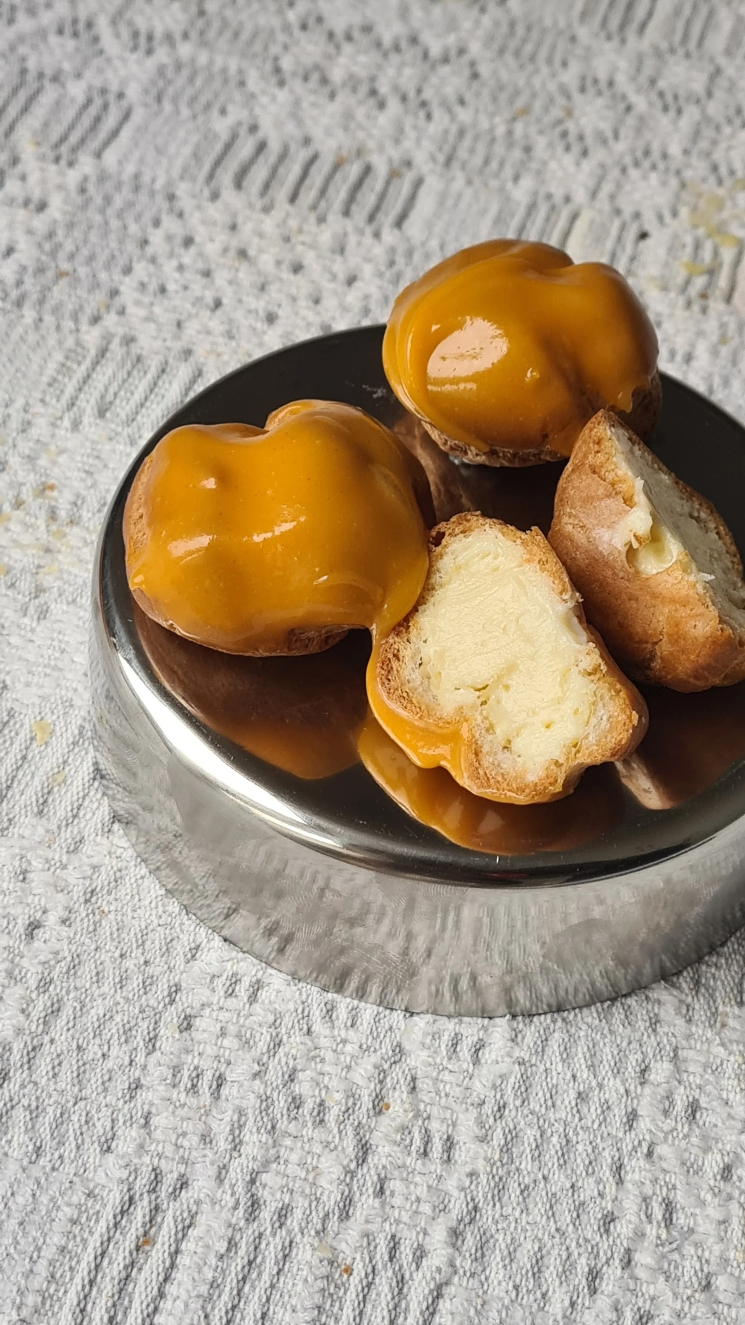 Two cream-filled profiteroles topped with caramel sauce on a black plate, resting on a textured white fabric surface.