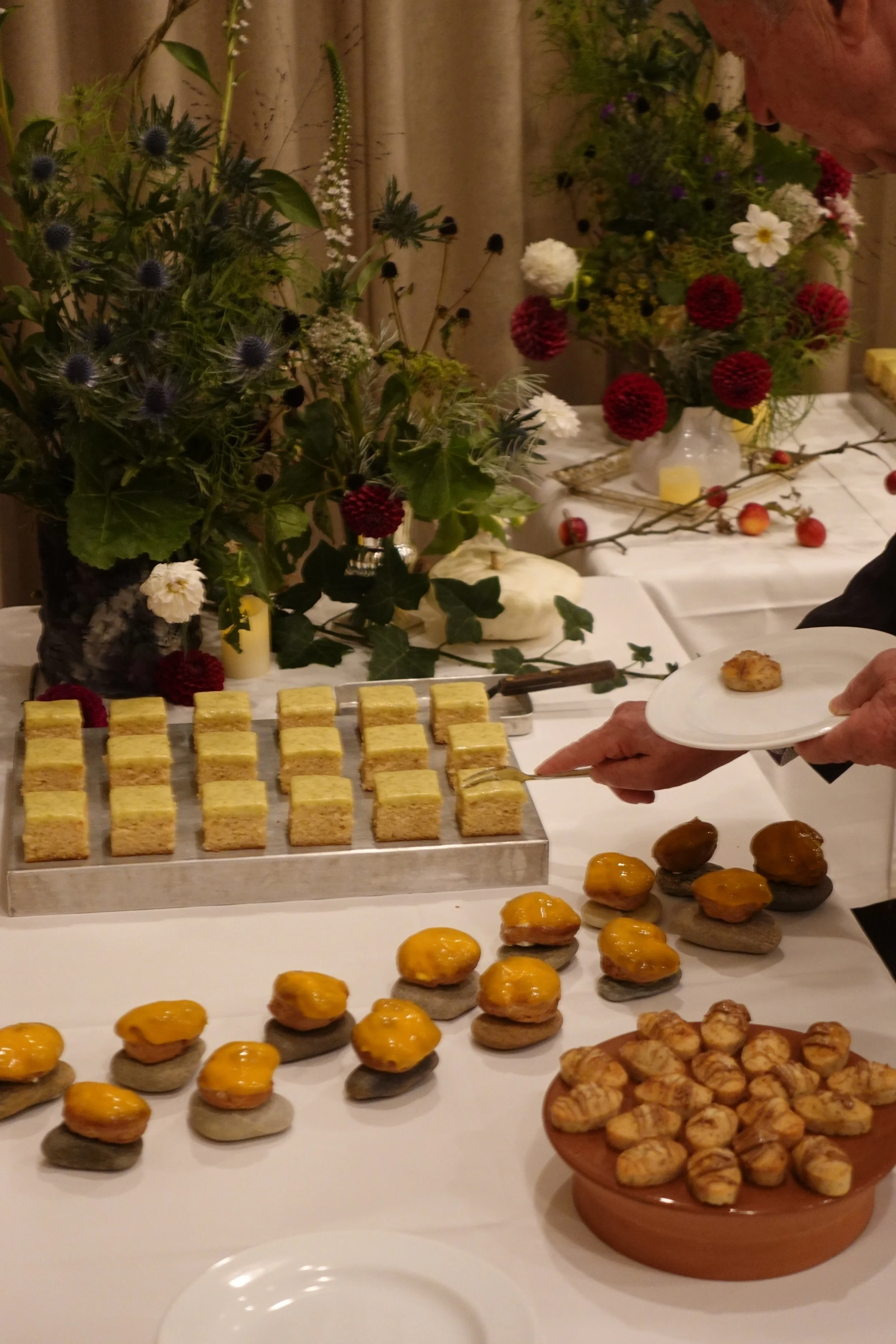 A person serving assorted appetizers, including topped crackers, small sandwiches, and a variety of finger foods, on a white table with floral arrangements in the background.