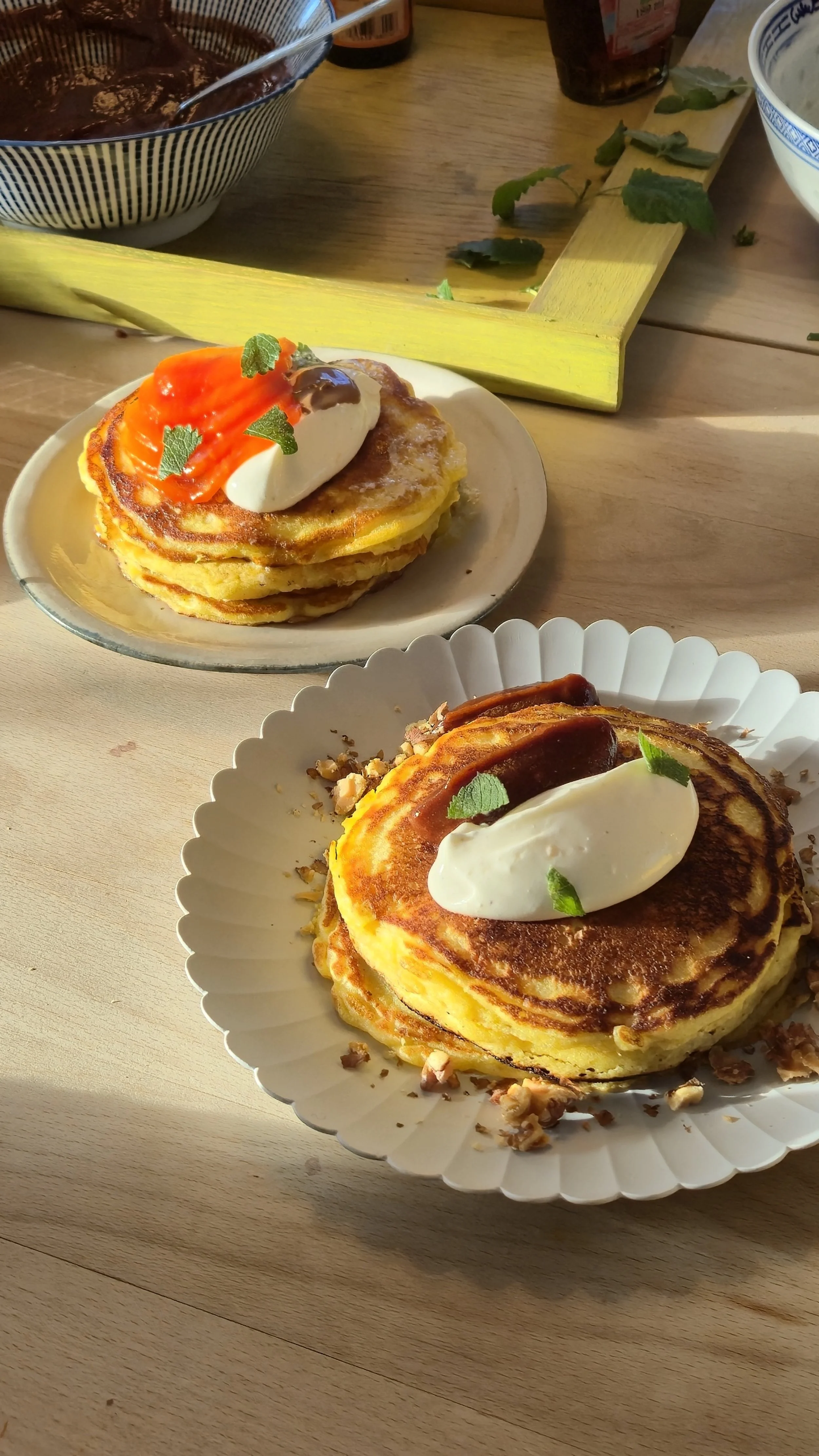 Stack of three golden-brown pancakes topped with whipped cream, caramel sauce, and mint leaves, served on a white paper plate, with another stack of pancakes topped with whipped cream, nuts, and syrup in the background.