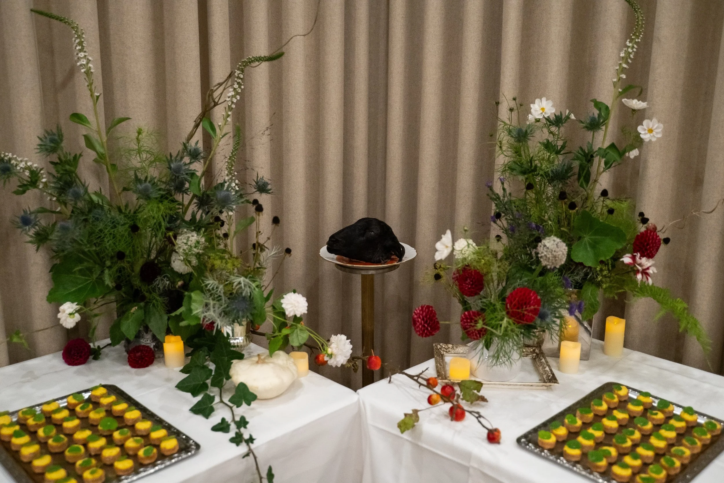 A funeral or memorial altar decorated with large floral arrangements, candles, and trays of small snack treats, with a black casket or body in the center.