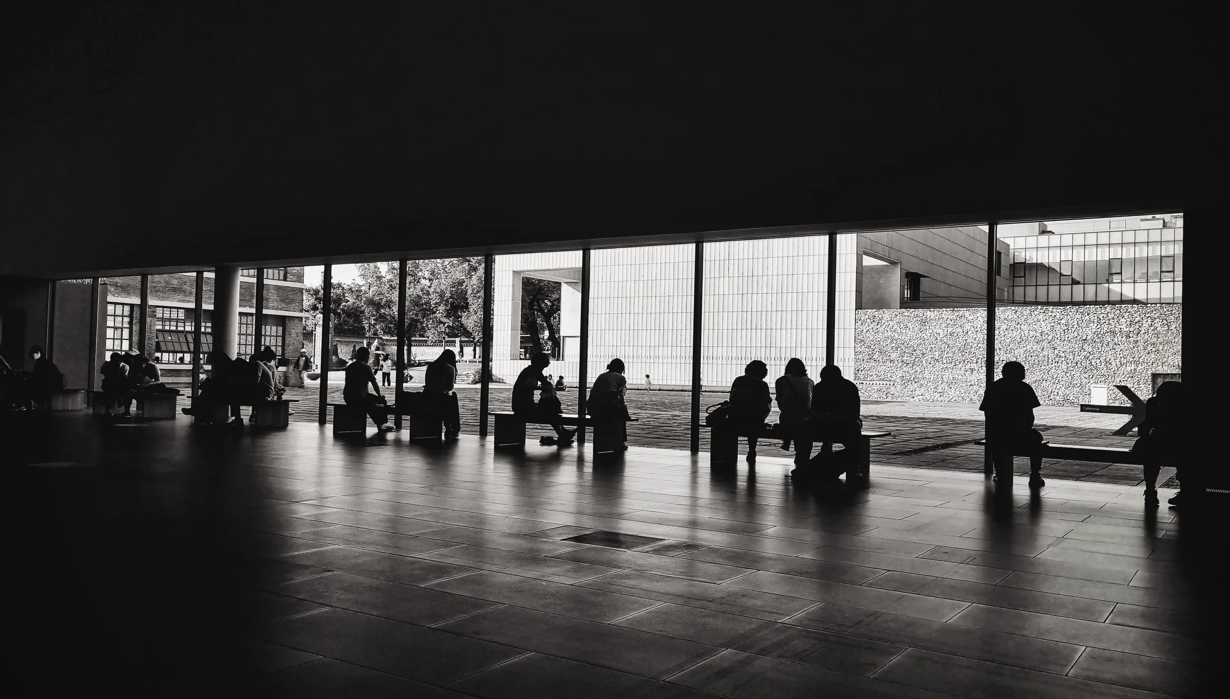 Silhouettes of people sitting on benches inside a modern building, with large windows showing an outdoor plaza with trees and modern architecture. Artistic investigation into the tension between historic and modern architecture in contemporary Seoul.