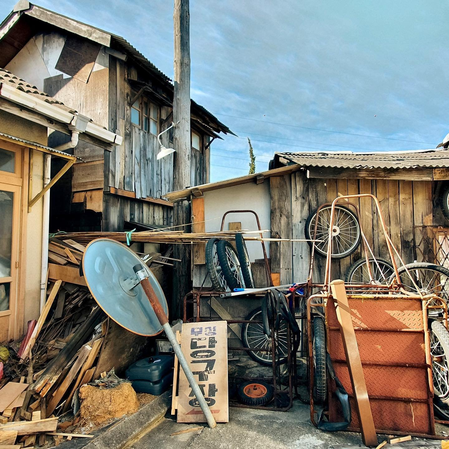 Cluttered yard with old bicycles, a rusty cart, a satellite dish, a sign reading 'Garage Sale,' and wooden scraps, all against weathered wooden structures and a cloudy blue sky.