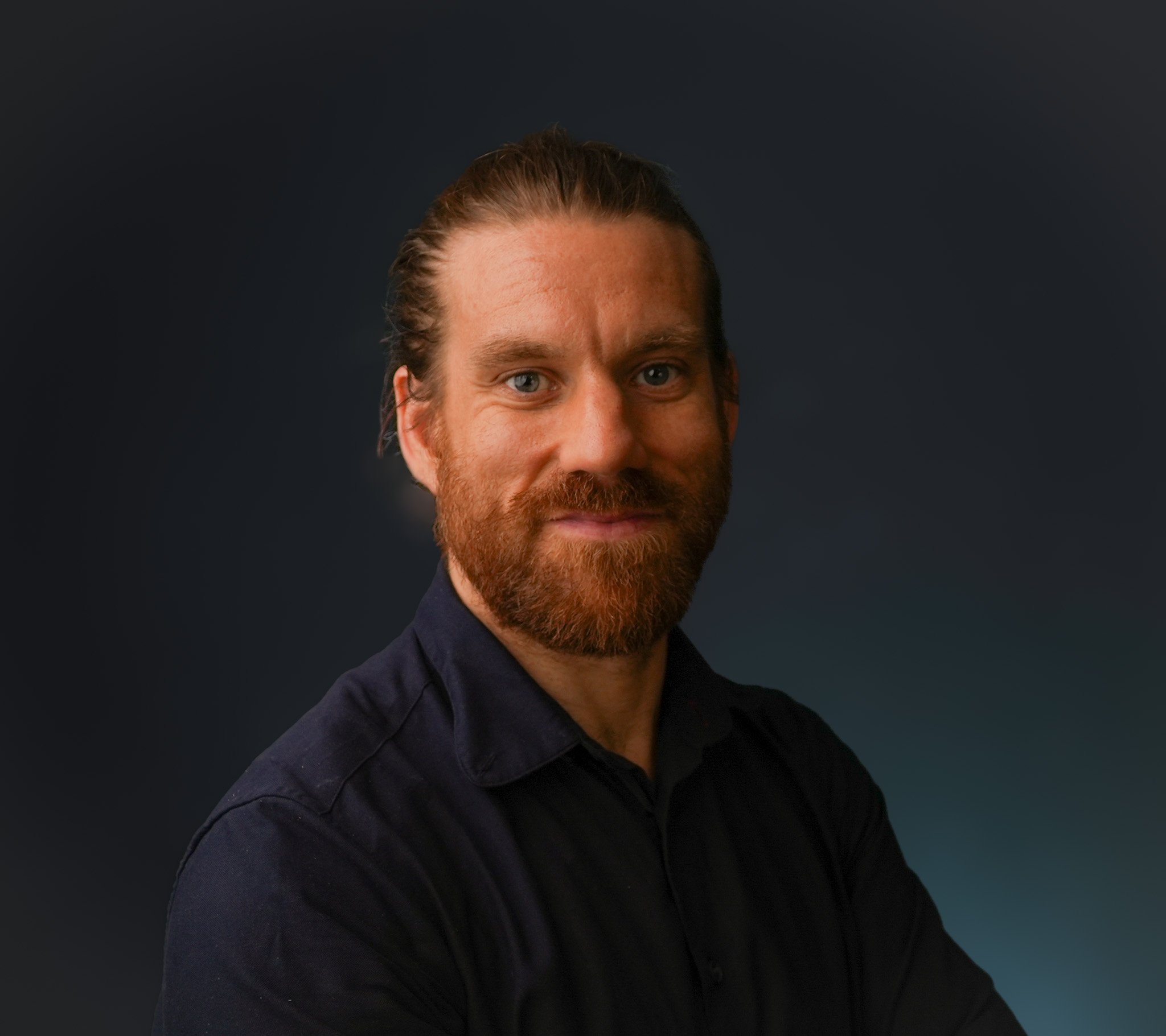 Portrait of a man with red hair and beard wearing a dark shirt, smiling against a dark background.