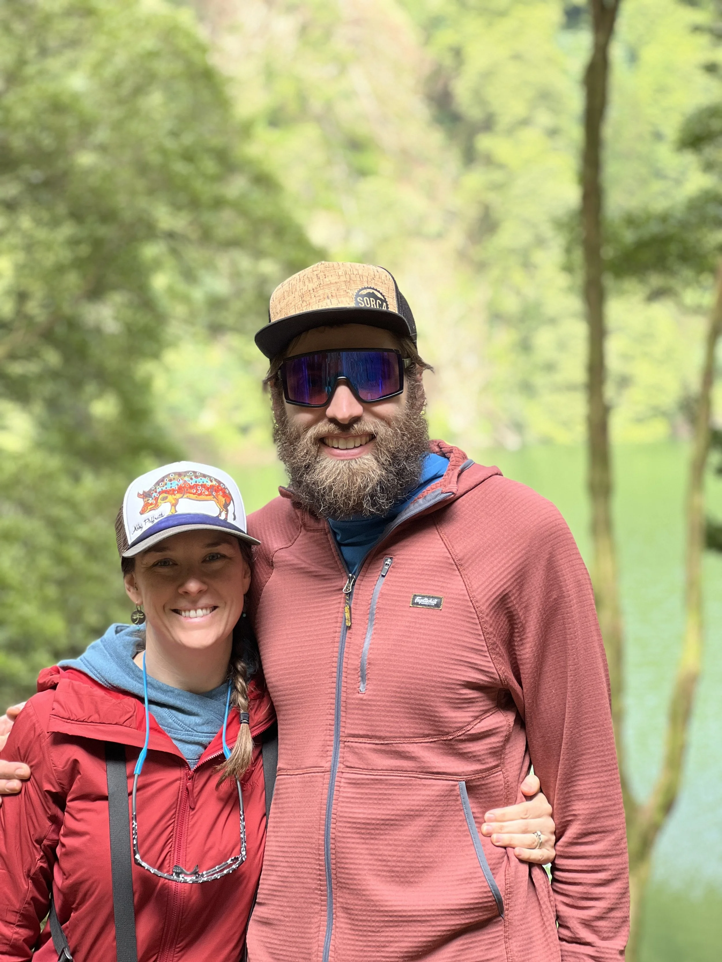A smiling couple standing outdoors in nature, with a background of green trees and a lake.