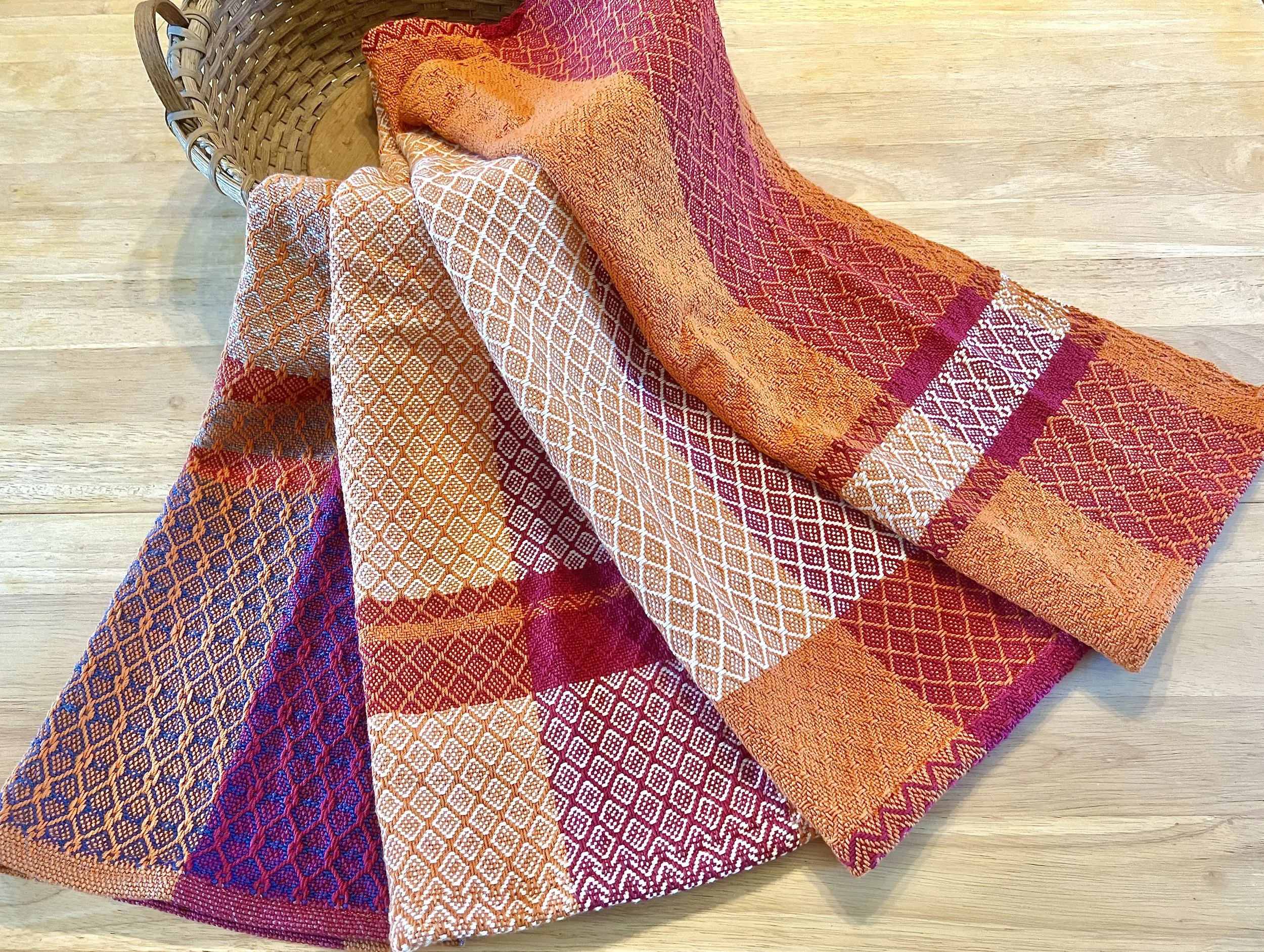 Colorful woven textiles (tea towels) with geometric patterns, partially draped over a basket on a wooden surface.