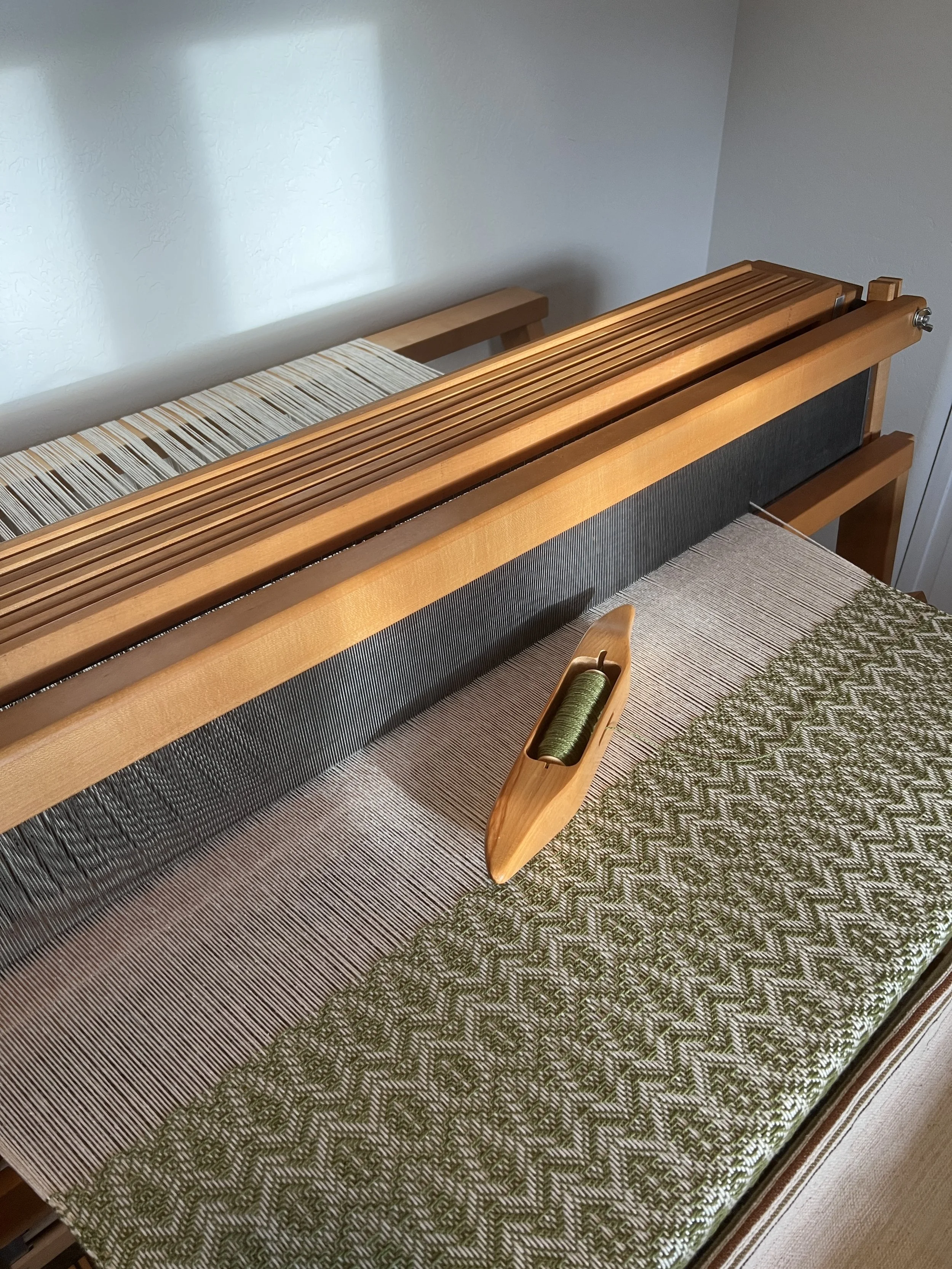 A wooden loom with a green patterned fabric and a small spool of green yarn in a wooden shuttle.