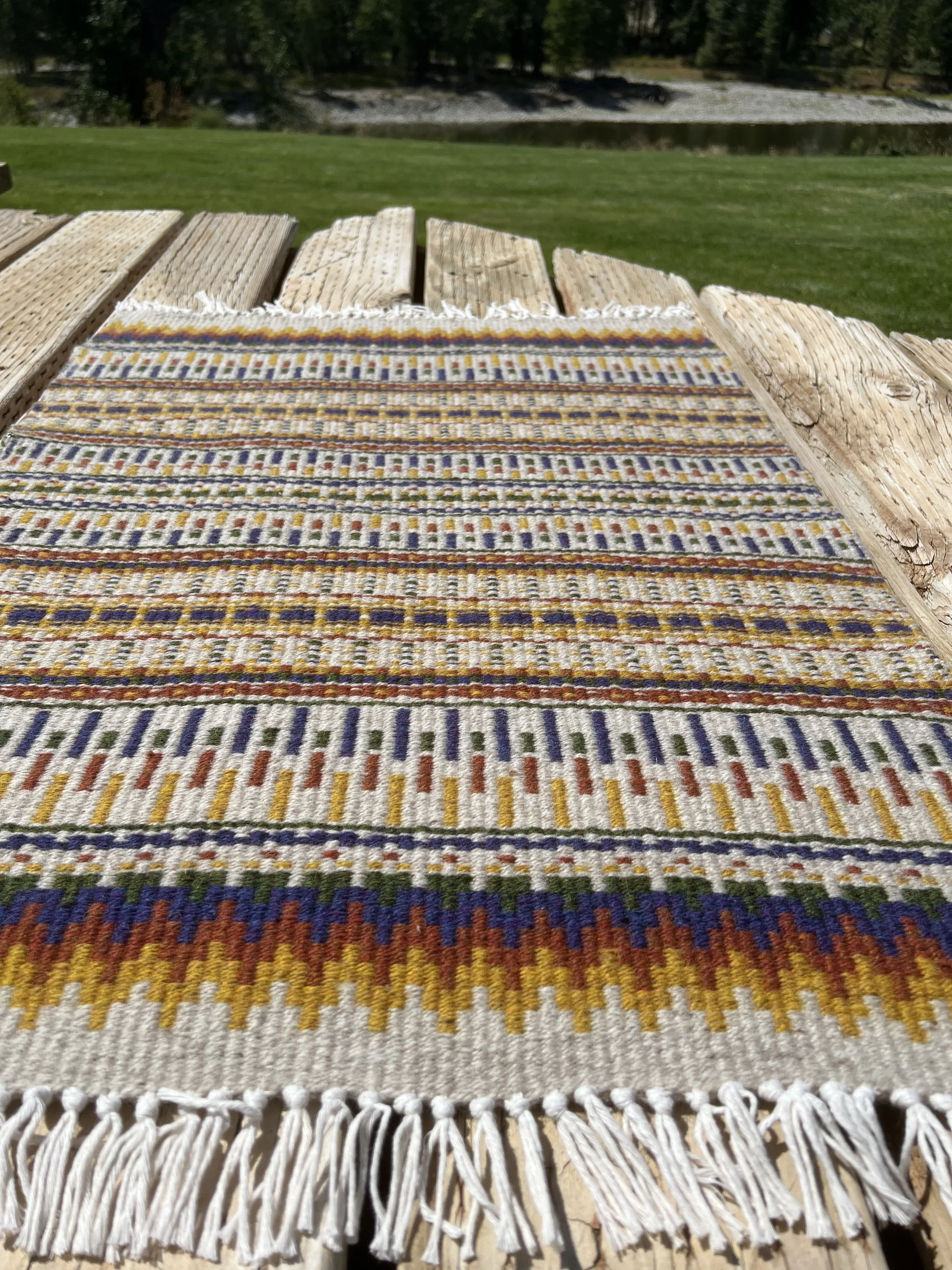 Colorful woven textile with fringe on a wooden bench outdoors, with grass, trees, and a body of water in the background.