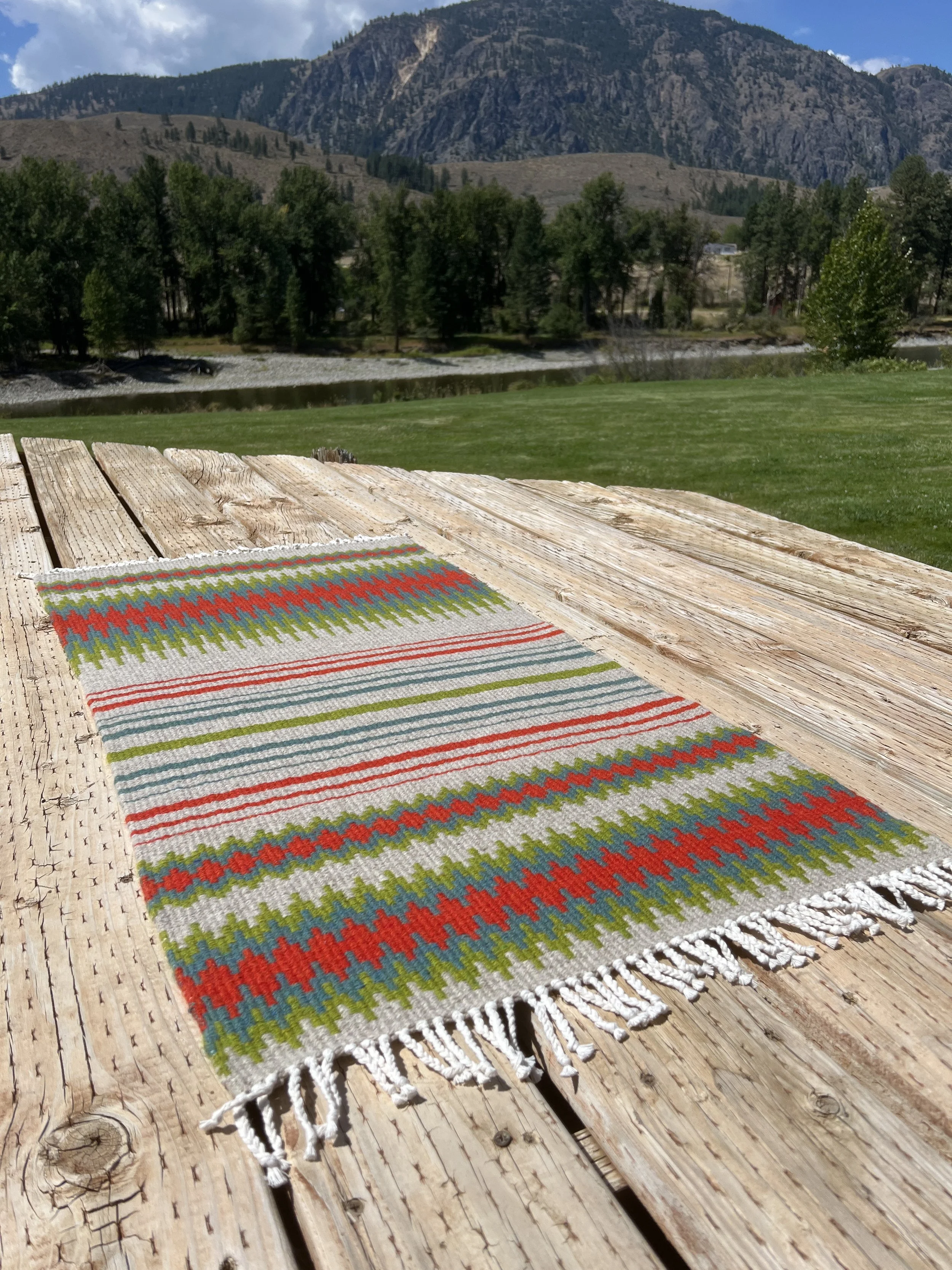 Colorful woven rug with red, green, blue, and white patterns draped over a rustic wooden deck, overlooking a grassy lawn, trees, a river, and mountains in the distance on a partly cloudy day.