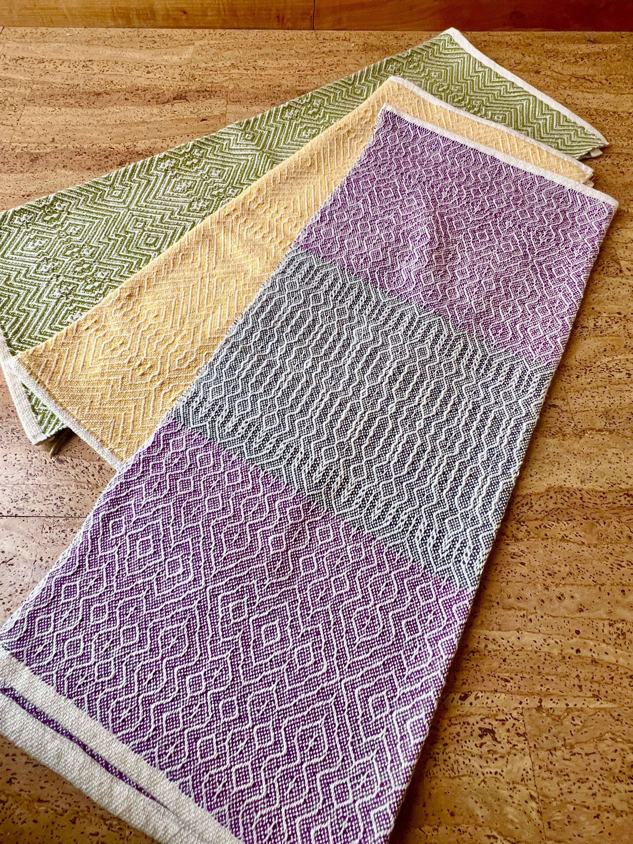 Three folded colorful dish towels with geometric patterns on a wooden floor.
