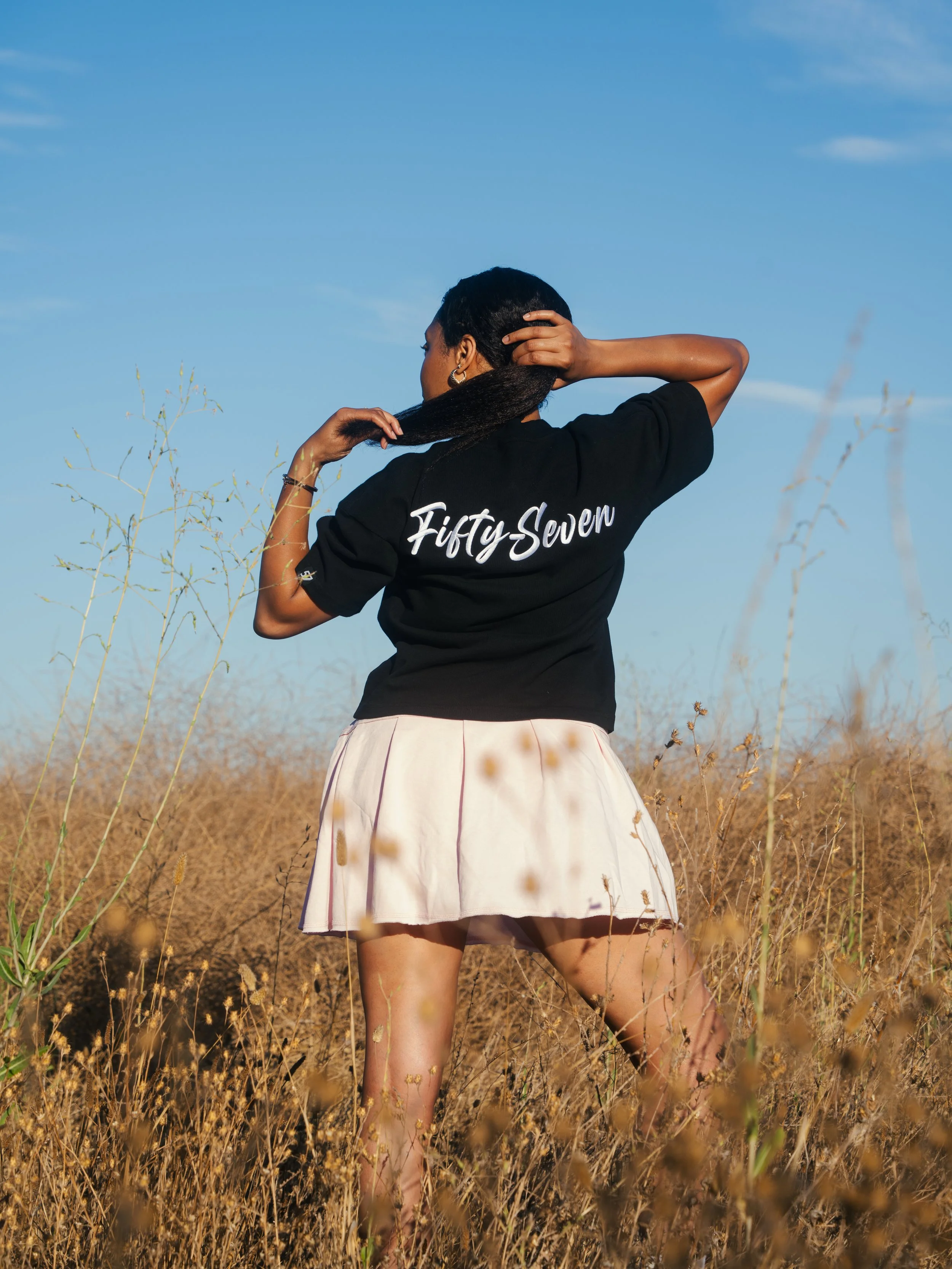 A woman with short black hair, wearing a black shirt with white text on the back, standing in a dry, grassy field under a blue sky with some scattered clouds.