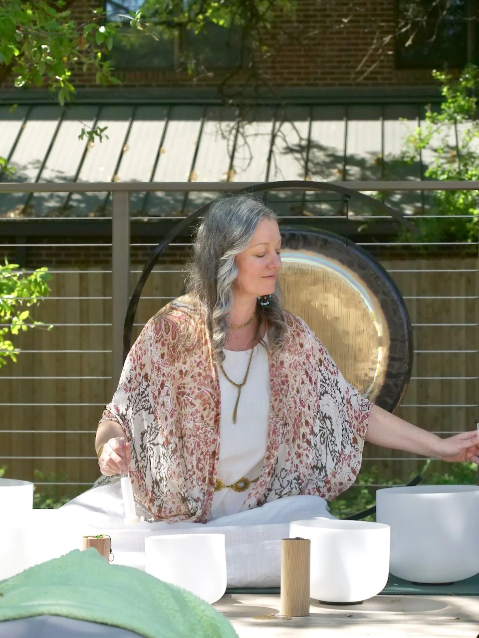 A woman with long, wavy gray hair playing crystal singing bowls outdoors on a sunny day.