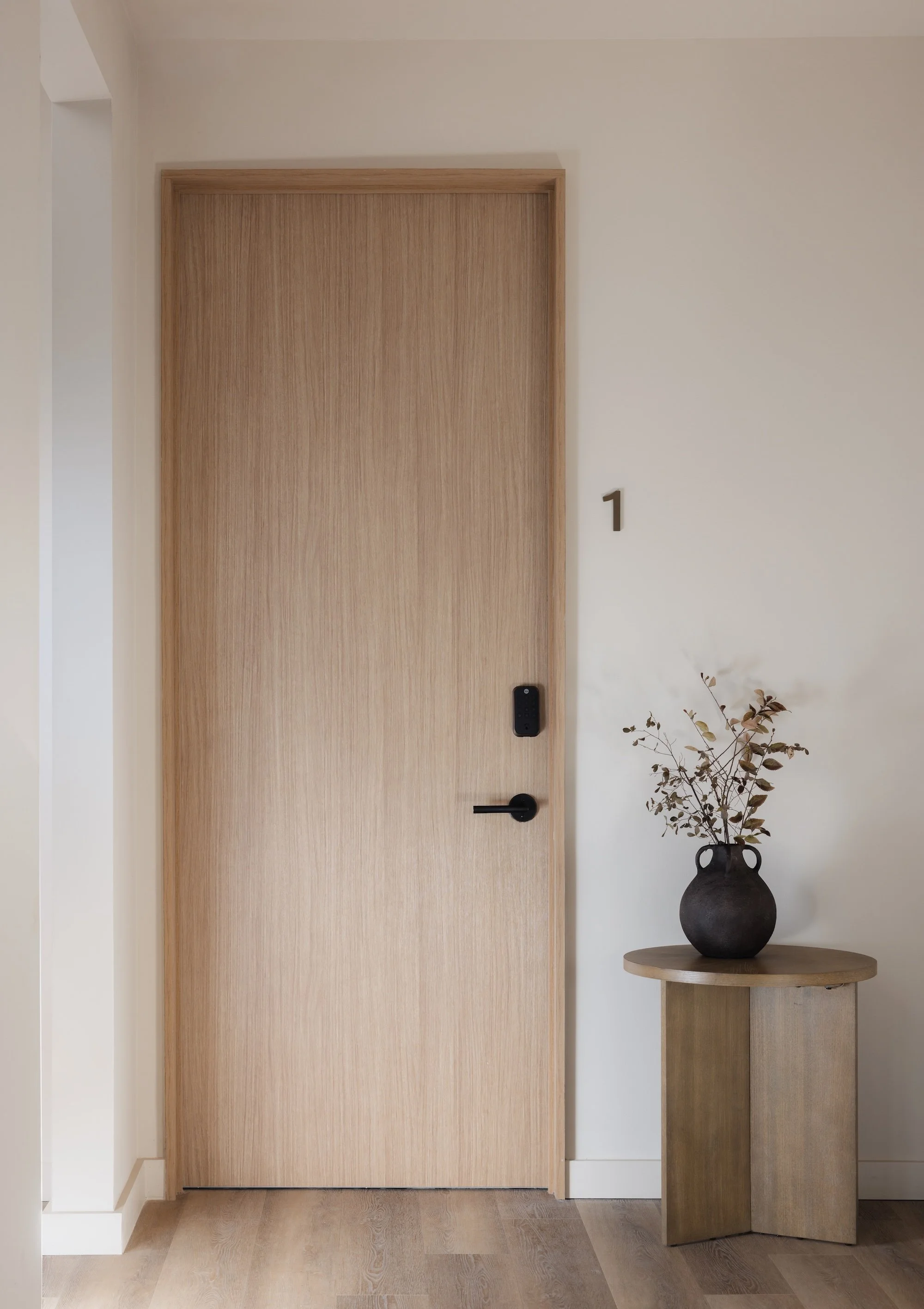 A minimalist interior scene featuring a closed wooden door with a black handle, a small side table to the right with a black vase holding dried branches, and a house number 1 on the wall.