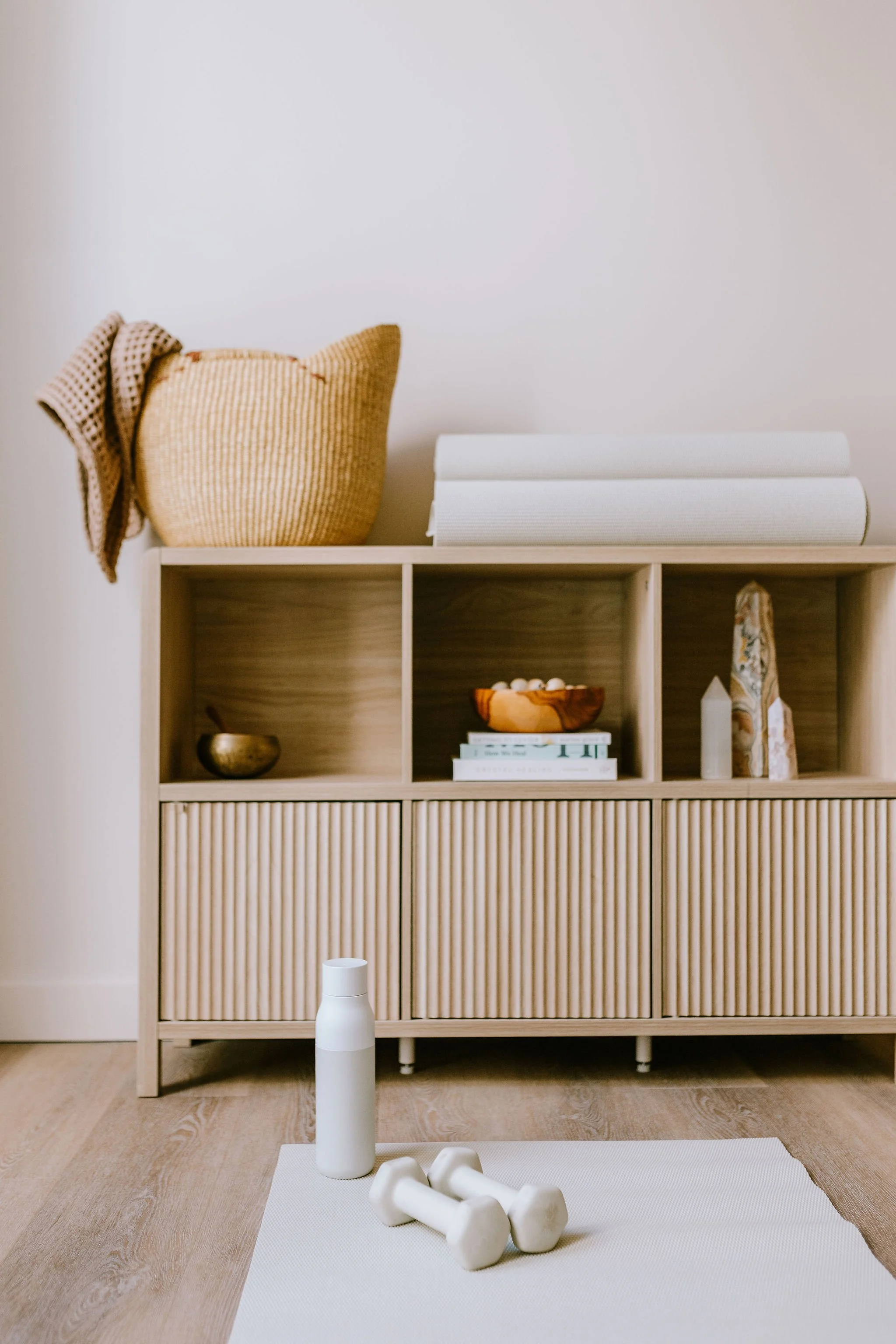 Minimalist room with a wooden shelf holding a woven basket, rolled yoga mats, books, and decorative objects. A white yoga mat is on the floor with white dumbbells and a water bottle on a mat nearby.