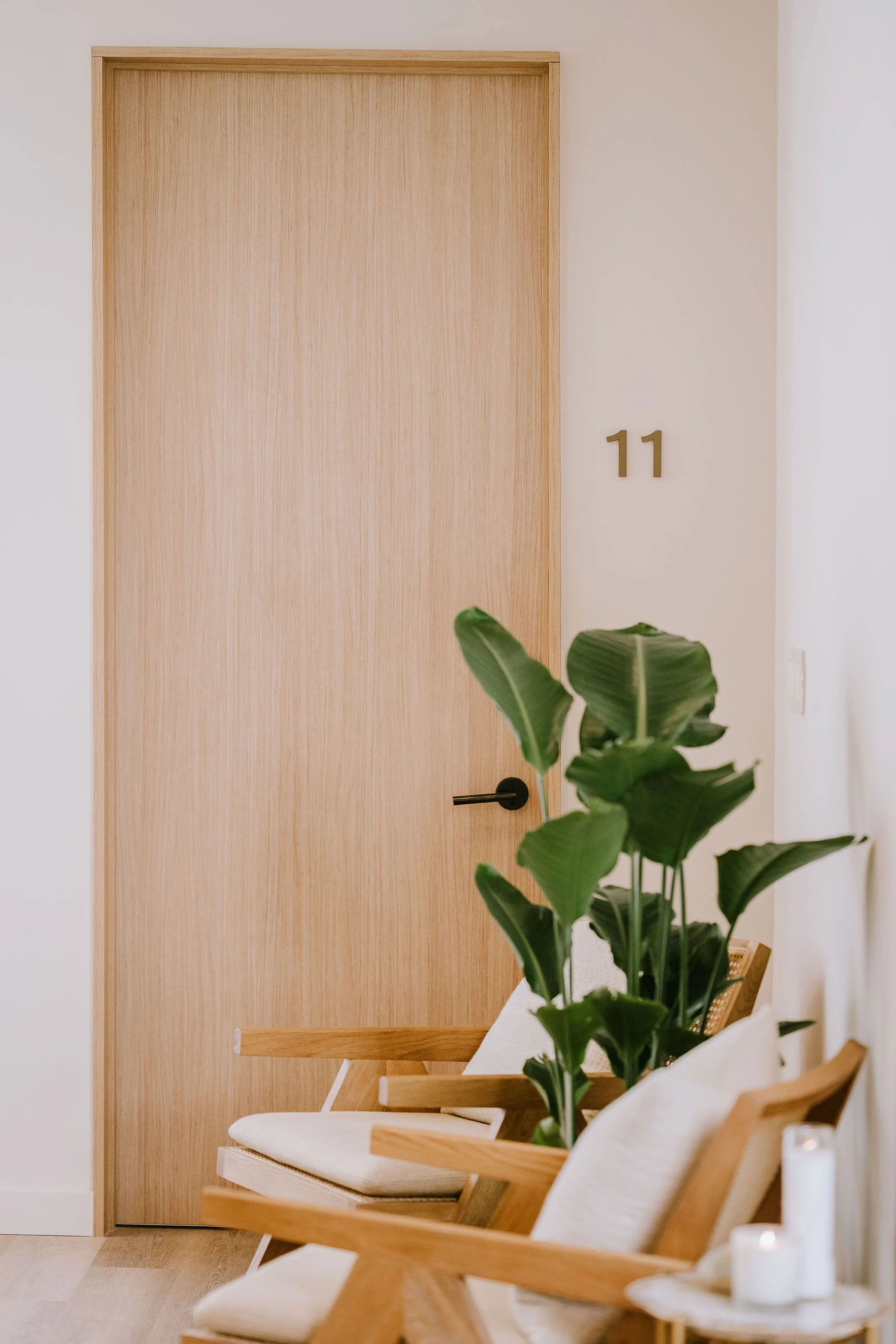 A wooden door with a gold house number 11 on a white wall. In front of the door, there are three wooden chairs with white cushions, and a tall green leafy plant partially obscures the view. Candles are on the tables.