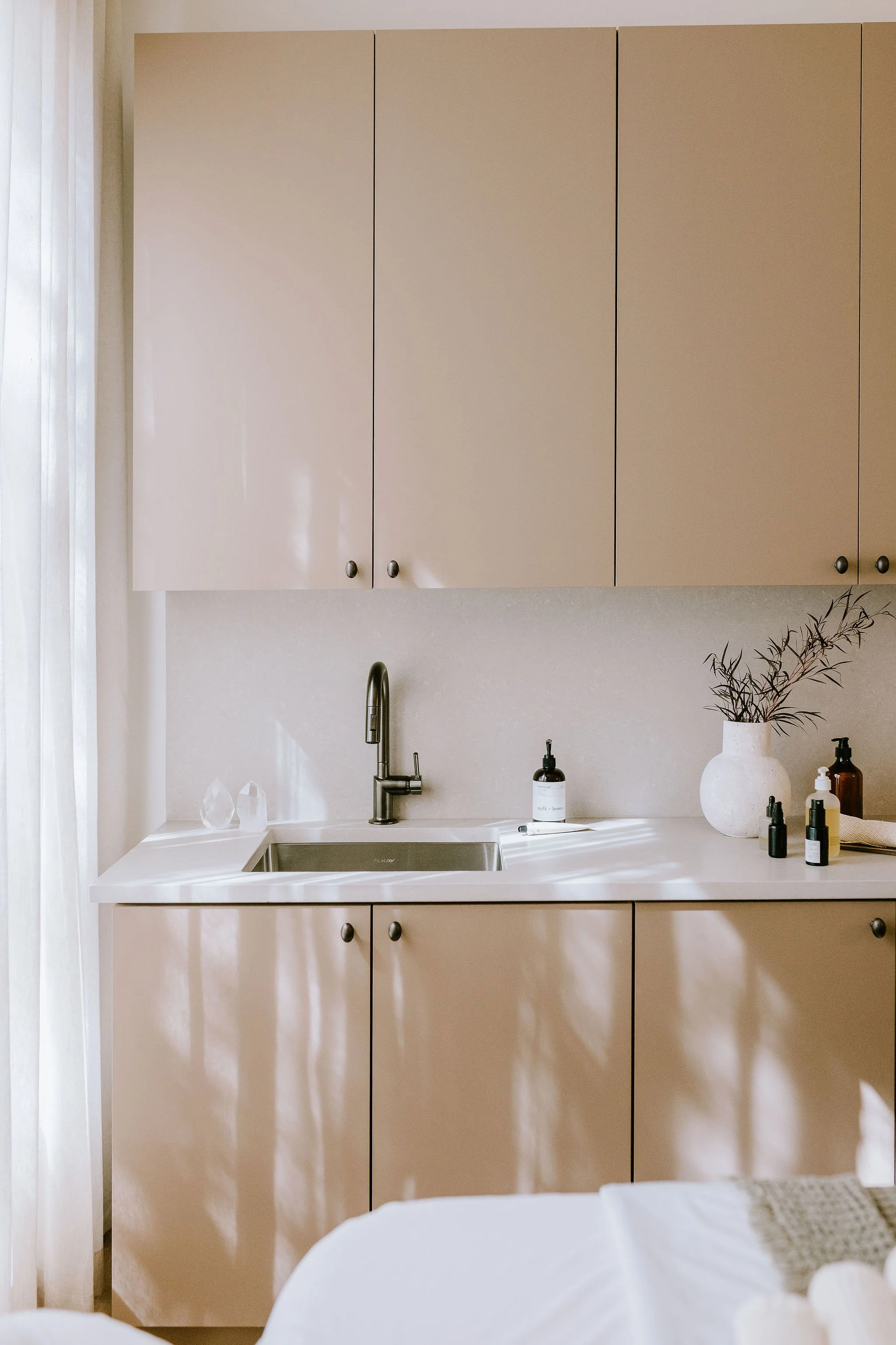 A minimalist kitchen with beige upper and lower cabinets, a stainless steel sink, a chrome faucet, and a white countertop. Decor includes a white vase with dried branches and several bottles and containers.