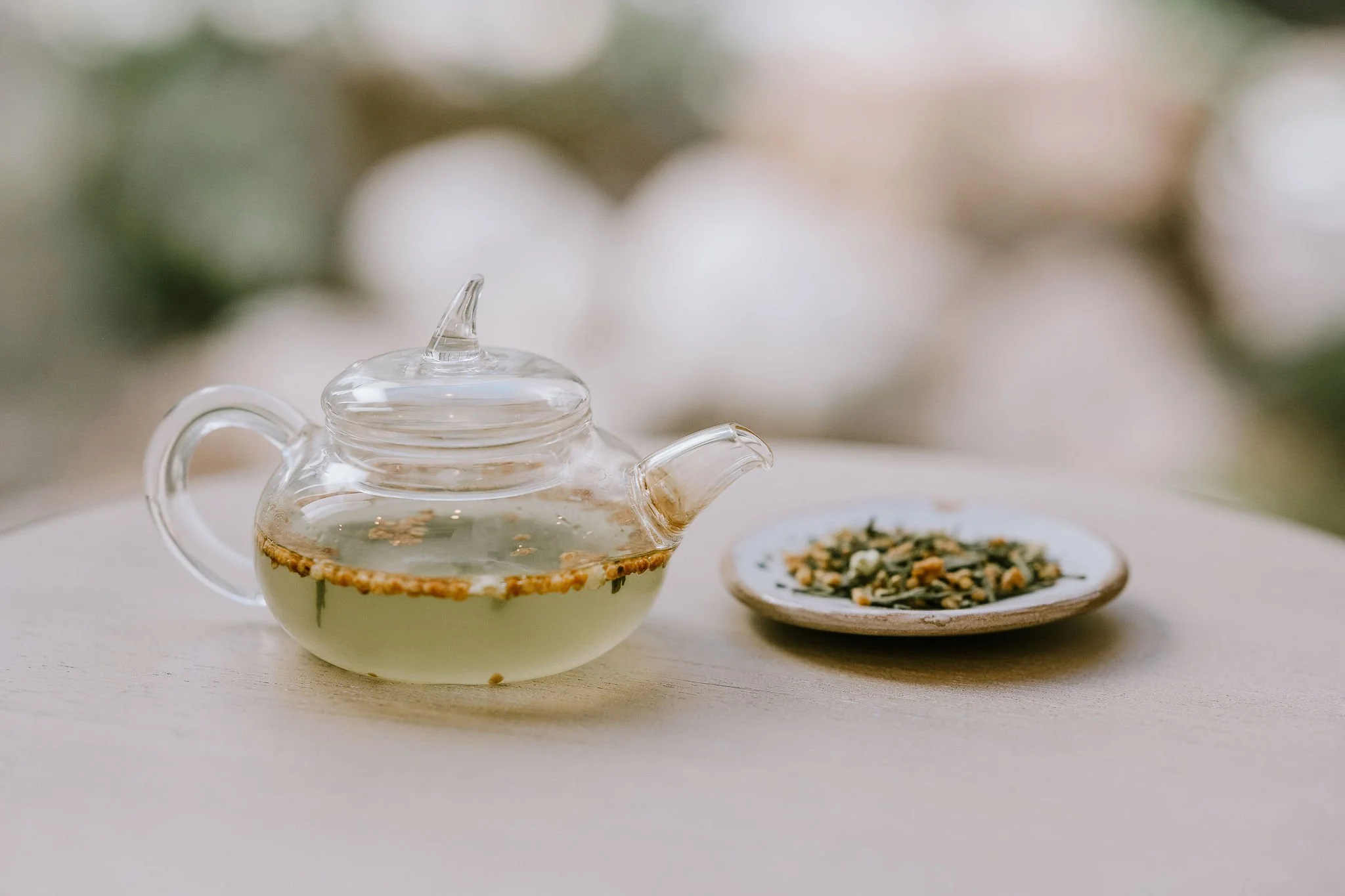 A glass teapot with a clear glass lid, filled with herbal tea, sitting on a white surface, with a small plate of dried herbs or tea leaves nearby blurred in the background.