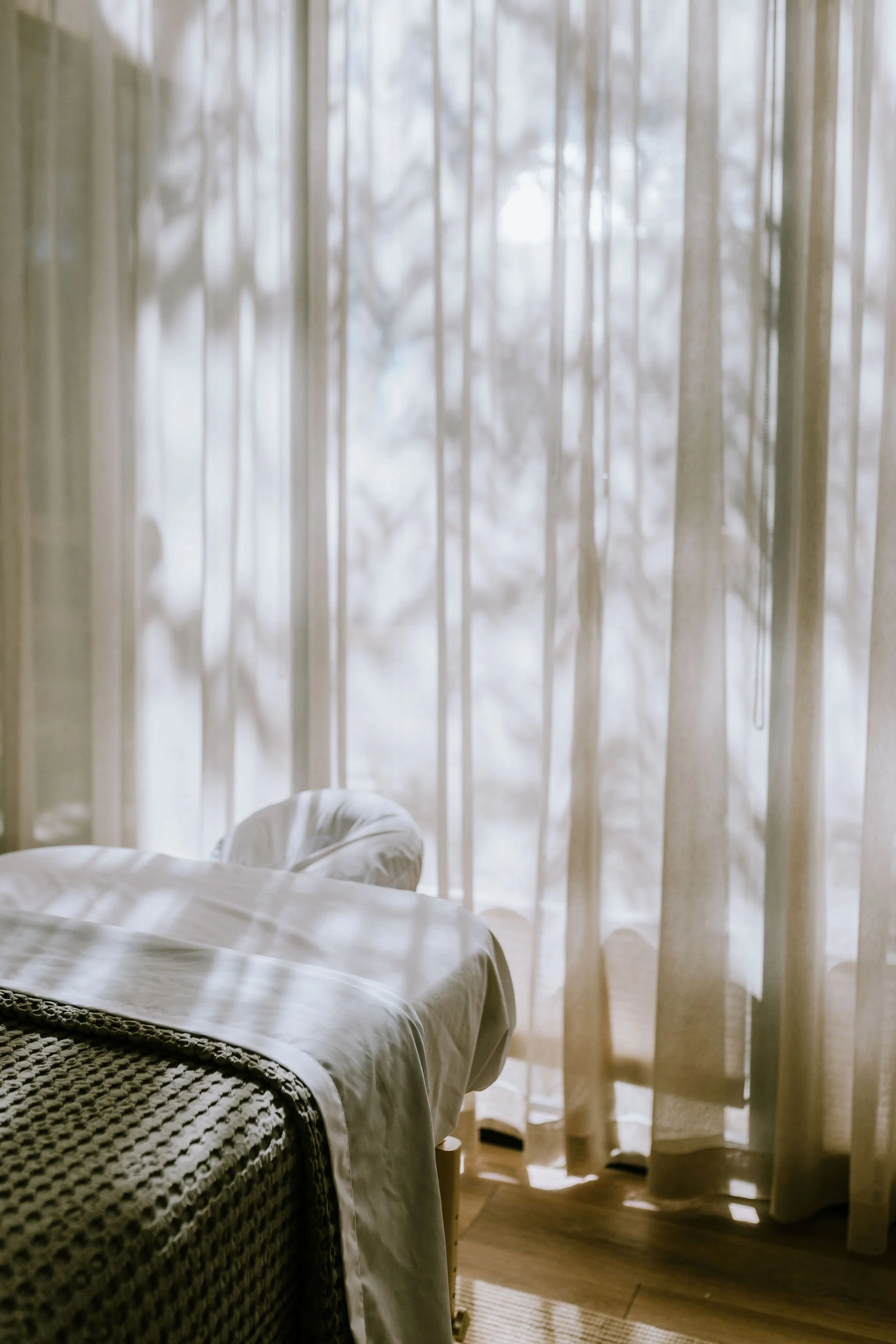 Sunlight filters through sheer curtains, illuminating a bed with white sheets and a textured blanket in a cozy room.