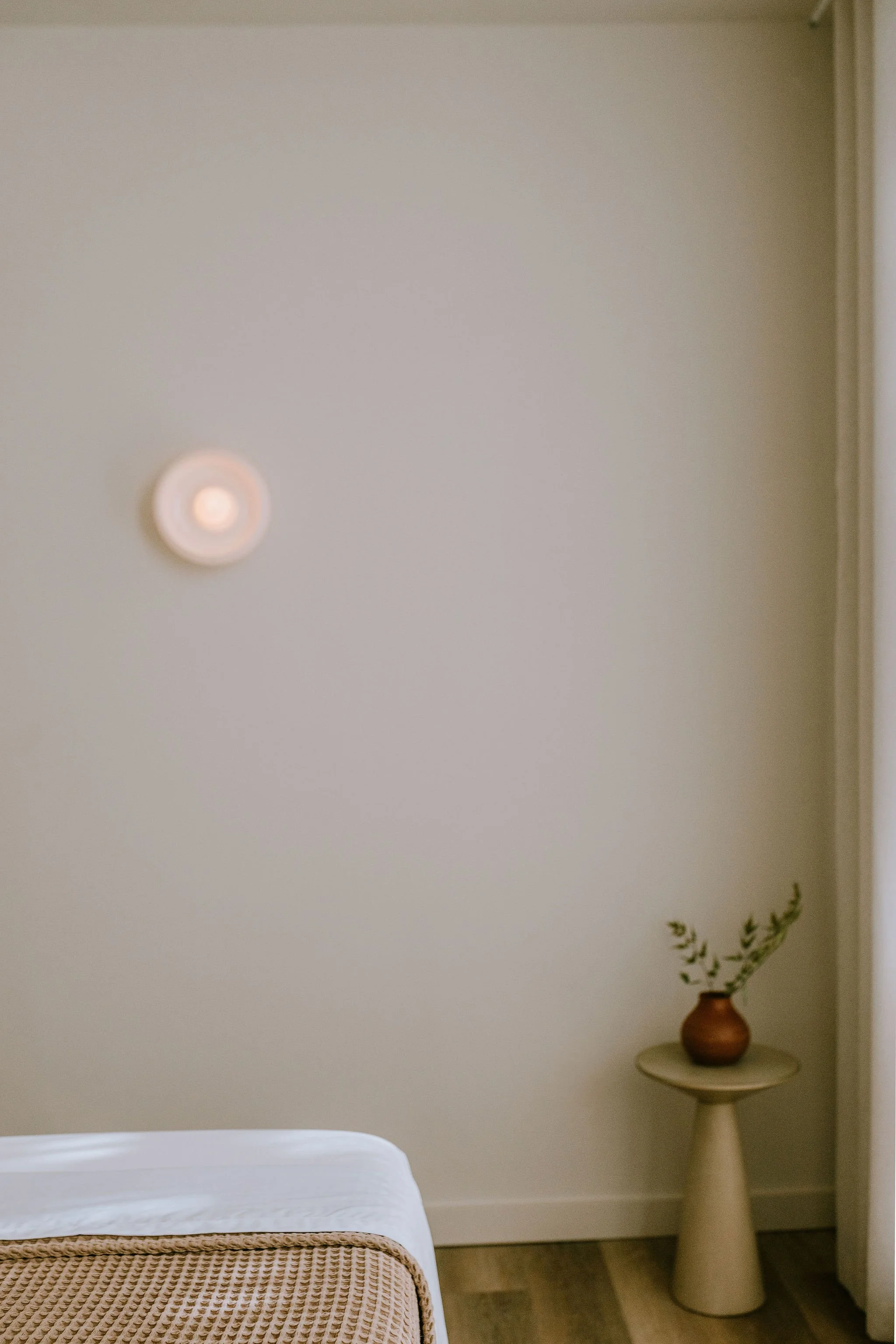A bedroom corner with a white bedspread, a woven blanket at the foot, a beige wall, a small metallic side table with a vase and plant, and a circular ceiling light.