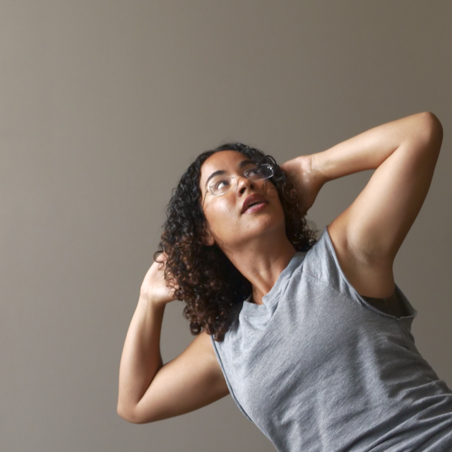 A woman with curly hair and glasses wearing a sleeveless gray shirt, posed against a plain background, with her hands behind her head and looking upward.