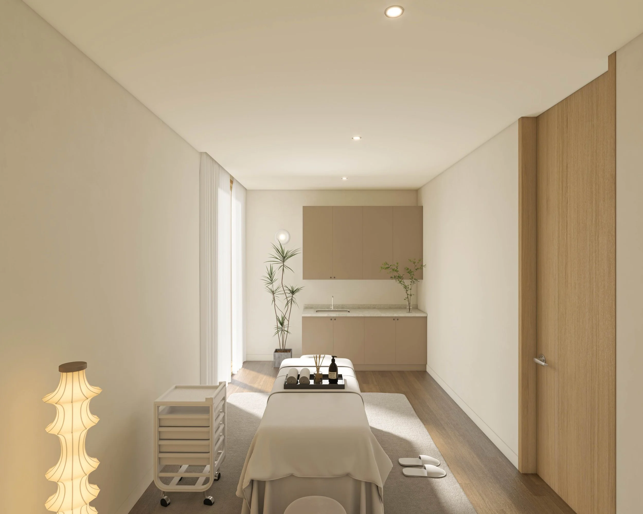 A minimalist massage therapy room with a massage table, a small rolling cart, a floor lamp, and a reception area with a small sink and cabinets, natural light coming through curtain-covered windows.