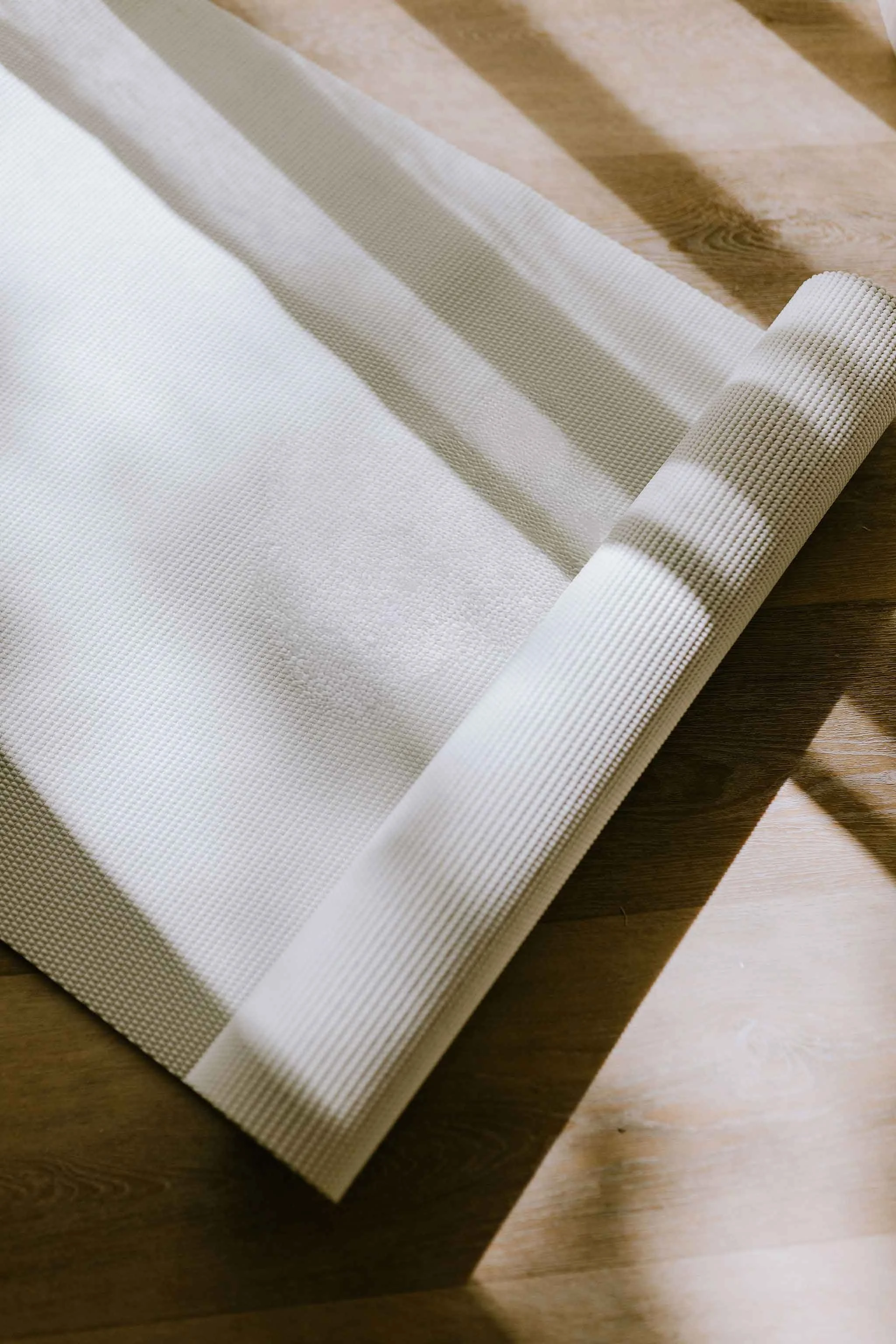 A partially unrolled white yoga mat with a textured surface, placed on a wooden floor, casting shadows.