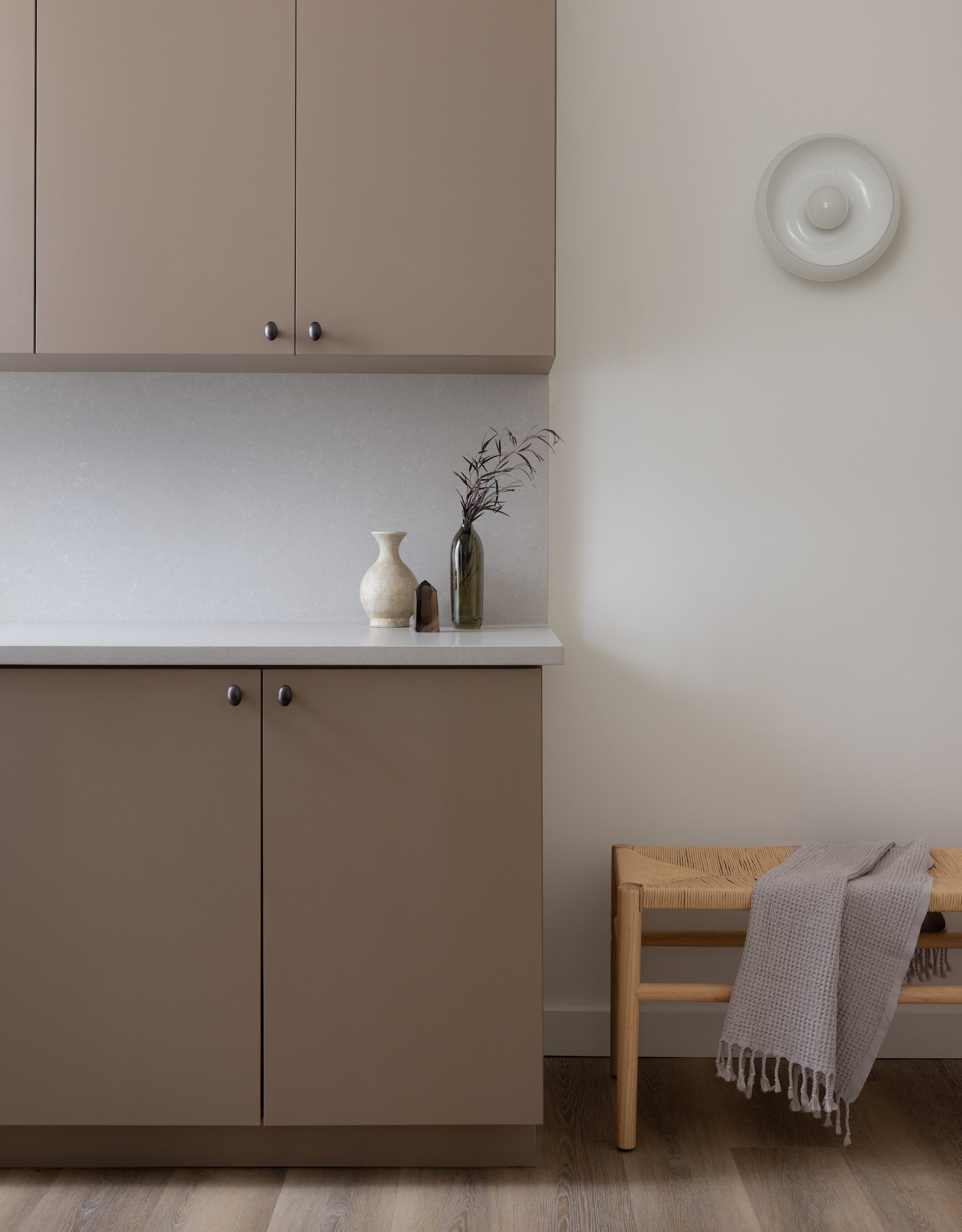 A beige kitchen cabinet with silver knobs, a white countertop, and a beige wall. On the countertop, there are three decorative vases and a dried plant. To the right, a wooden bench with a gray, textured throw blanket draped over it.