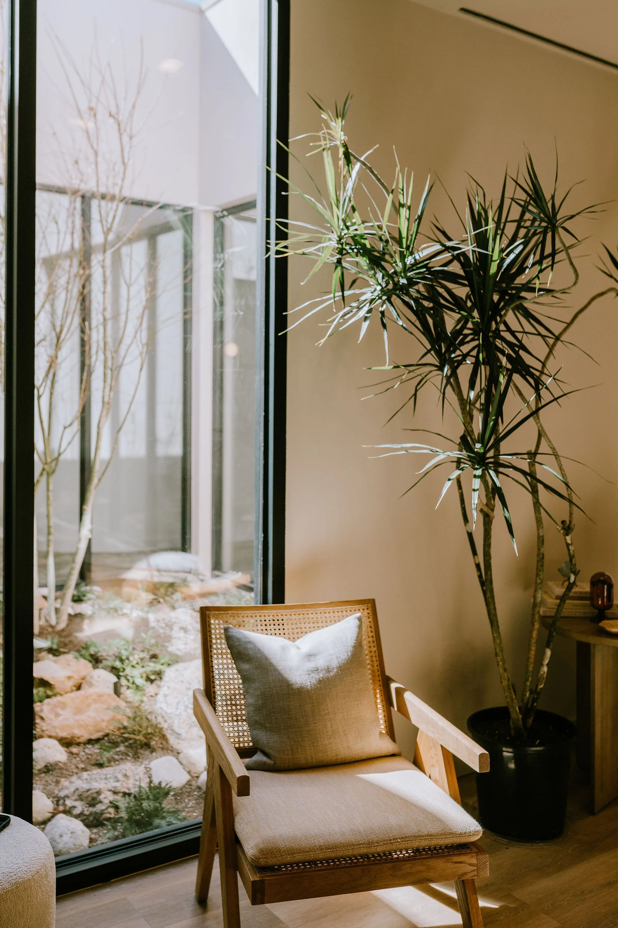 A cozy indoor space with a wooden armchair featuring a beige cushion and a gray pillow, next to a large potted plant with long, narrow leaves. There's a sliding glass door leading to a small outdoor garden area with rocks and plants.