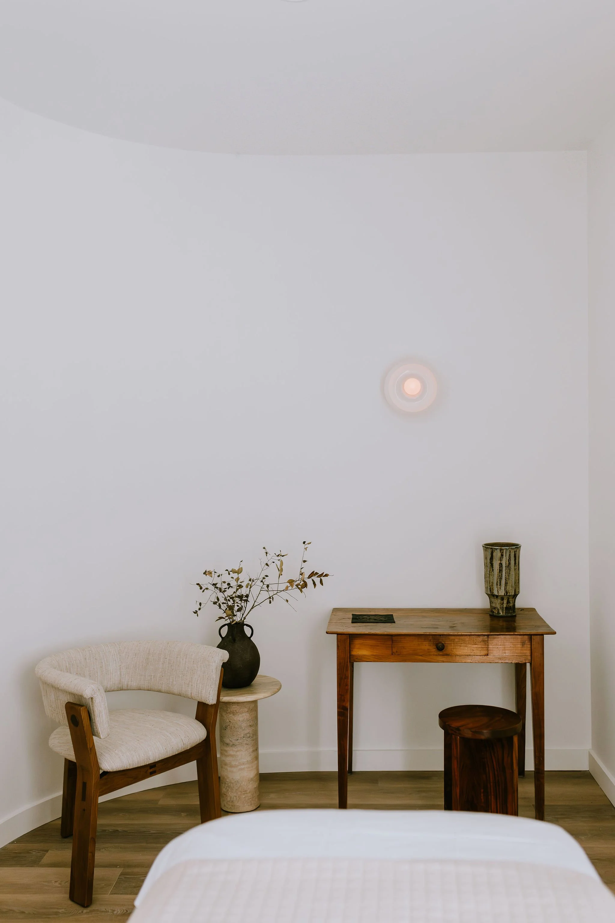 A minimalistic bedroom corner with a white wall, a wooden table, a patterned vase with a tall wooden vase, a smaller dark wooden stool, a beige armchair with a cushion, a small black rectangular object on the table, and some dried foliage in a dark vase.