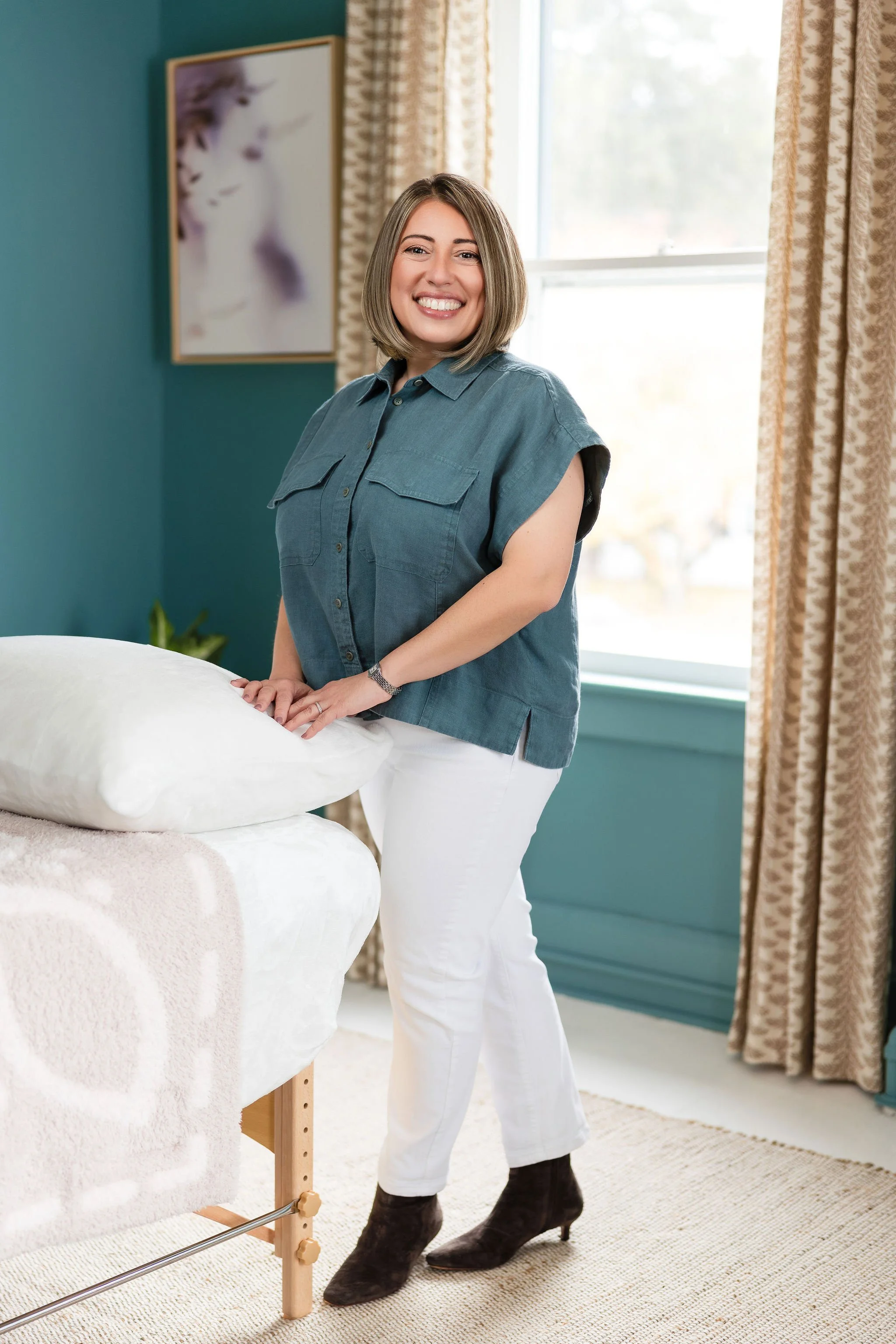 A woman with shoulder-length light brown hair smiling, standing beside a bed in a cozy, well-lit room with teal walls, beige curtains, and abstract artwork on the wall.