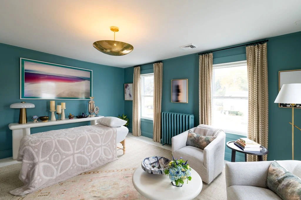 A cozy living room with teal walls, beige curtains, a white bench, two white armchairs, a round white coffee table with a flower arrangement, a dark side table with books, and a standing white lamp. Artwork decorates the walls, and sunlight streams through the windows.