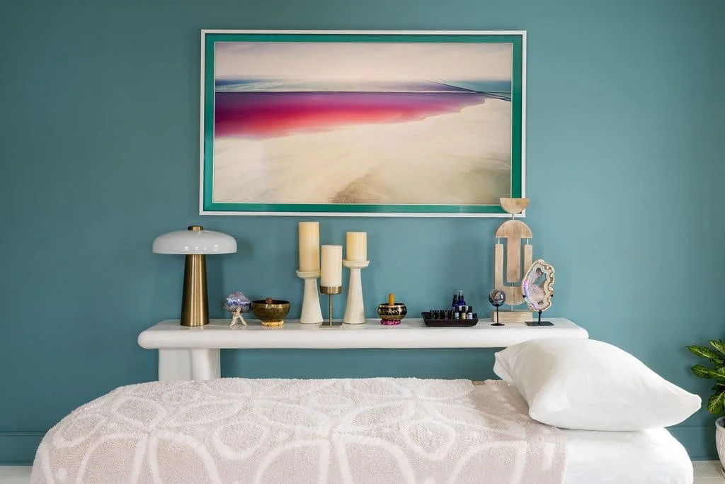 A bedroom with a teal wall, a white bed with a textured blanket, and a white nightstand holding decorative items including a lamp, candles, and abstract art pieces. A large landscape painting hangs above the nightstand.