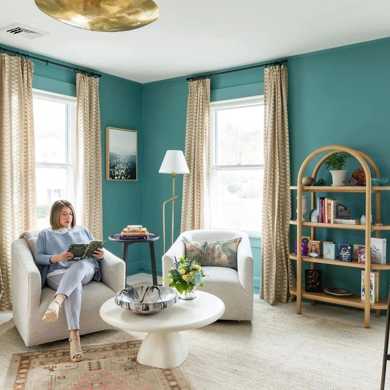 Living room with teal walls, beige curtains, two white armchairs, a black side table with books, a standing lamp, a round coffee table with a flower arrangement, and a wooden bookshelf with decorative items and books. A woman is sitting on one of the armchairs reading a book.