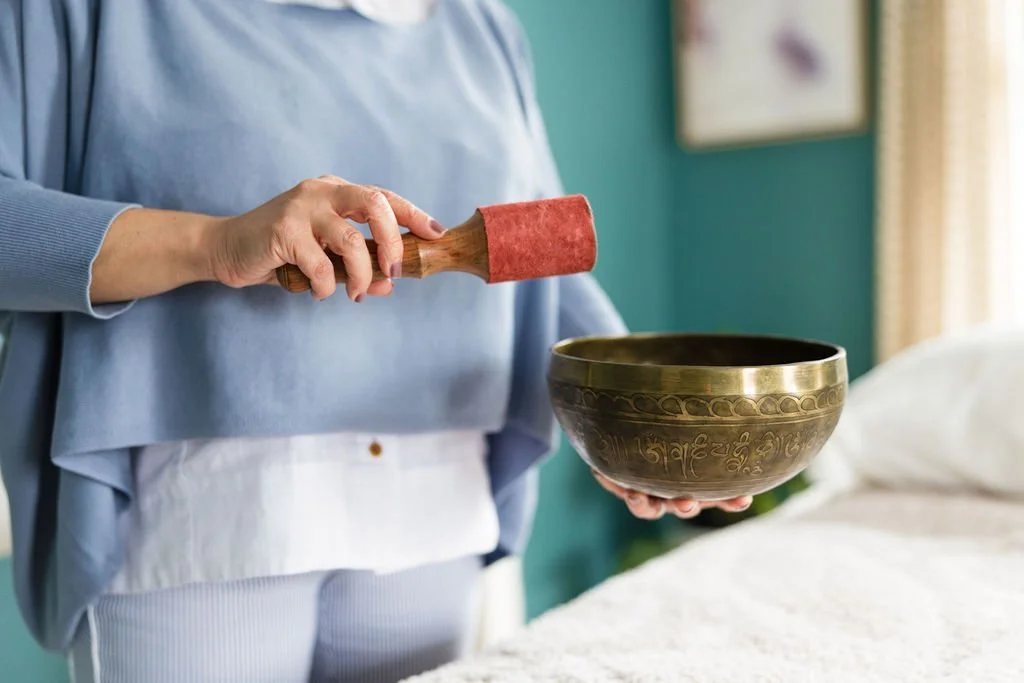 Person holding a Tibetan singing bowl with a mallet in a room with teal walls and a bed in the background.