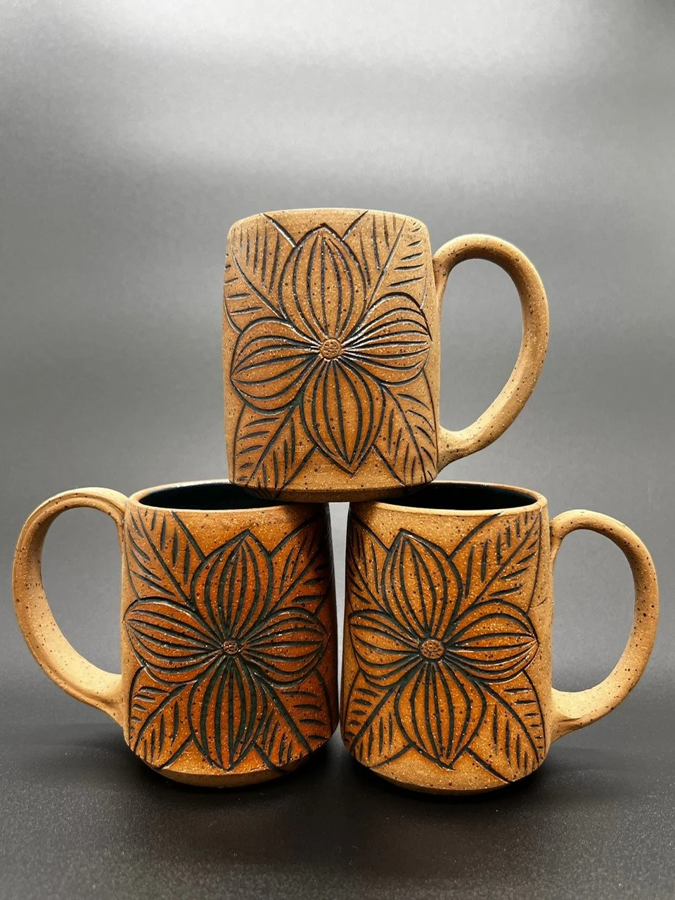 Three ceramic mugs with orange and black floral patterns, stacked with one mug on top of two mugs against a gray background.