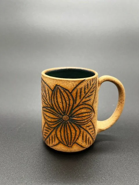 Ceramic mug with a large flower design on the front, placed on a dark surface against a plain gray background.