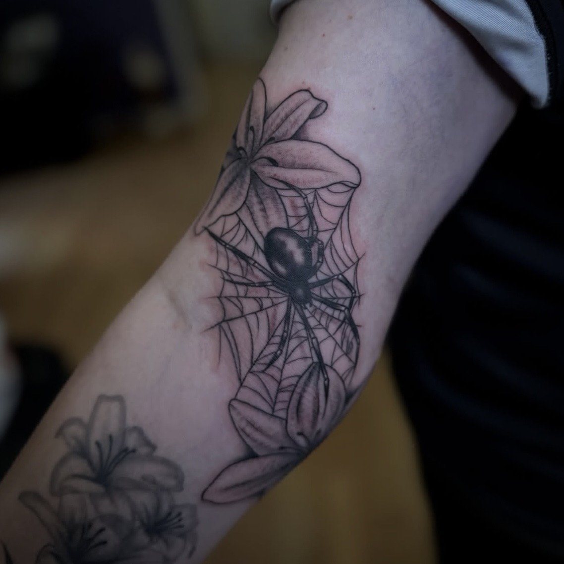 Tattoo of a spider with lily flowers and a spider web on a person's arm.