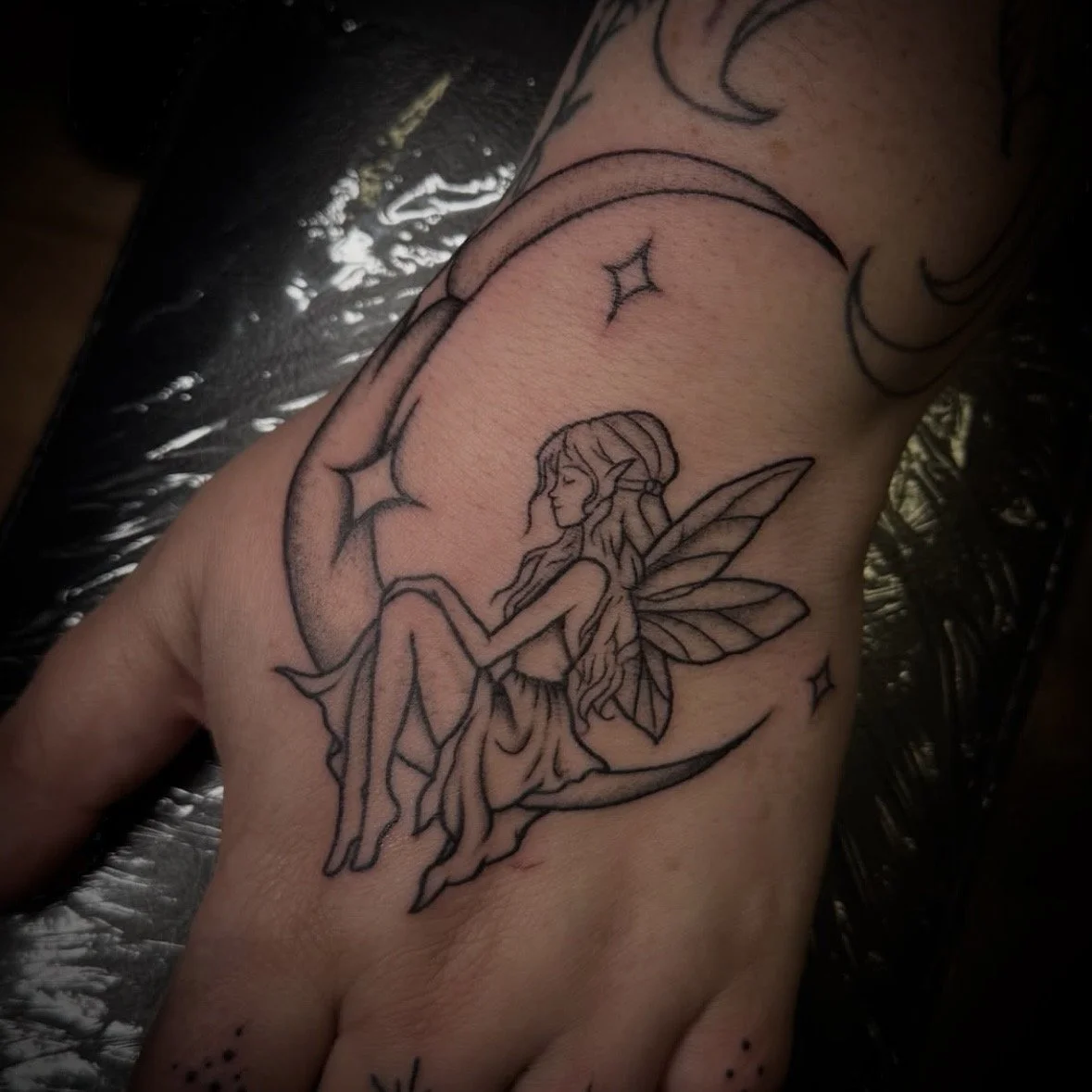Tattoo of a fairy sitting on a crescent moon with stars around, on a person's hand.