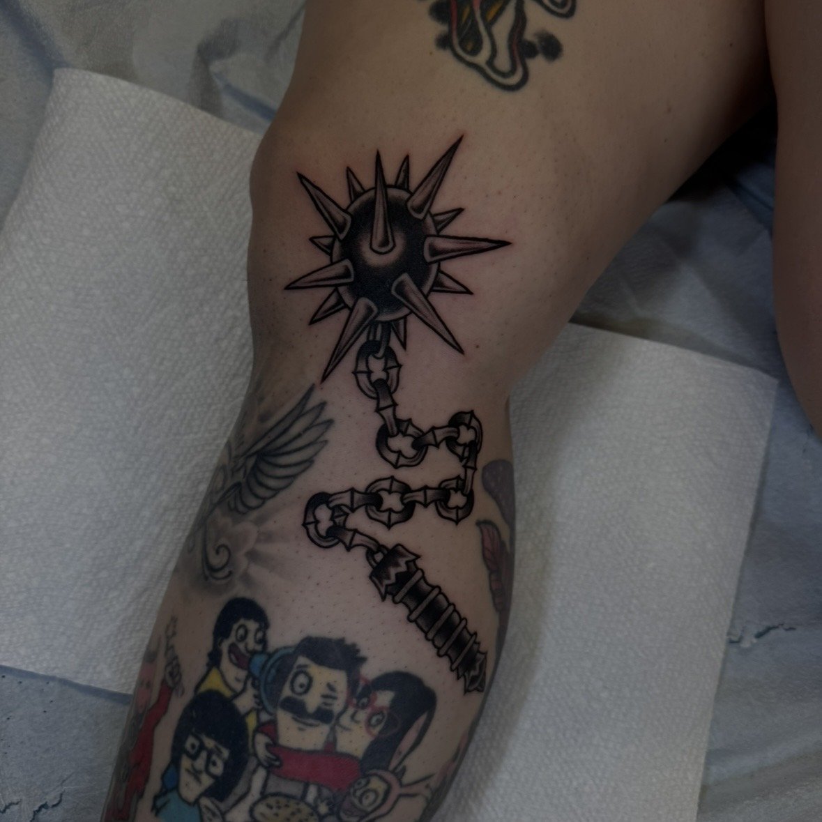 Tattoo on a person's leg featuring a black and gray spiked ball with a chain attached, and a cartoon character below.