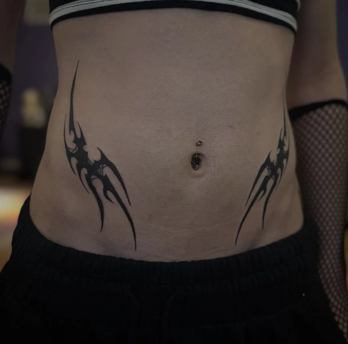 Close-up of a person's lower abdomen with black tribal tattoos on each side, a belly button piercing, and the person wearing black pants and fishnet sleeves.
