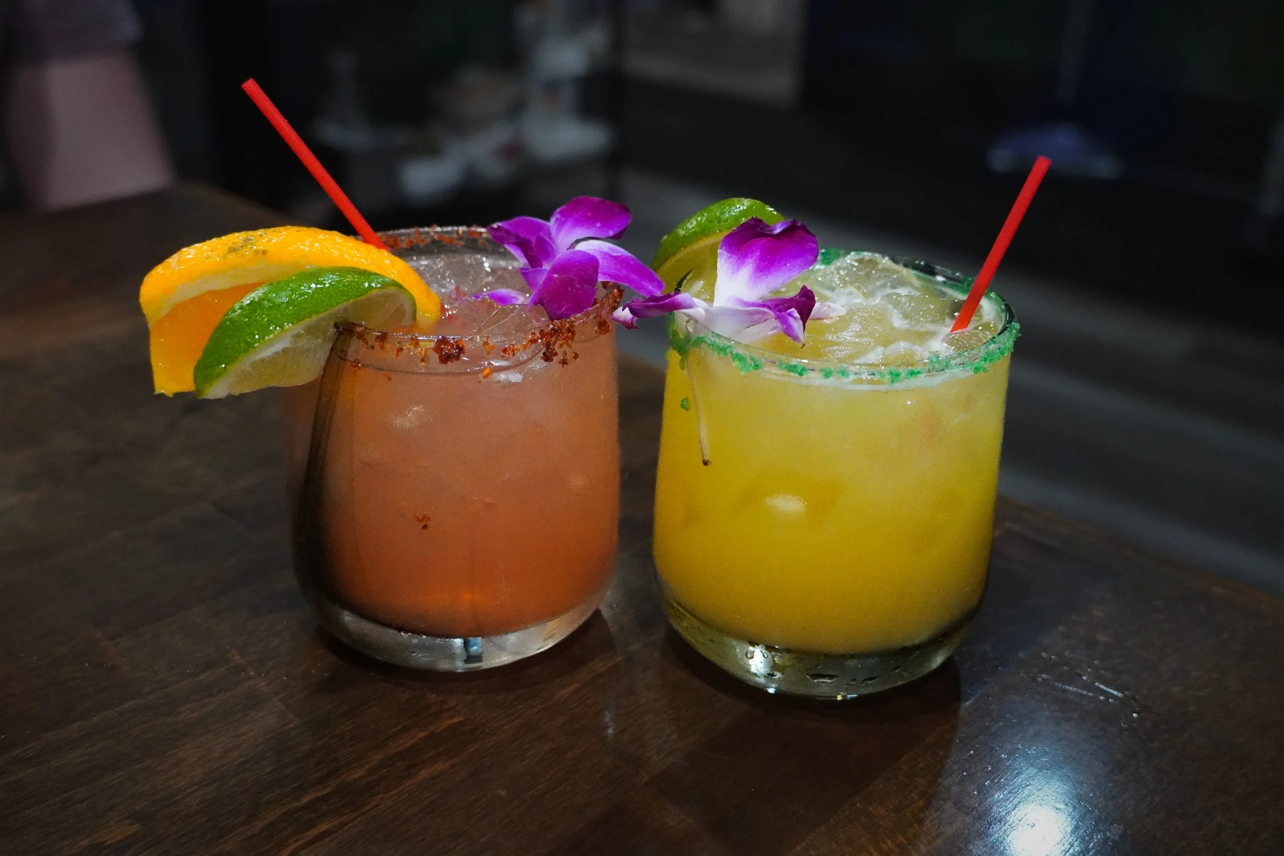 Two colorful cocktails on a wooden table, garnished with fruit slices and flowers.