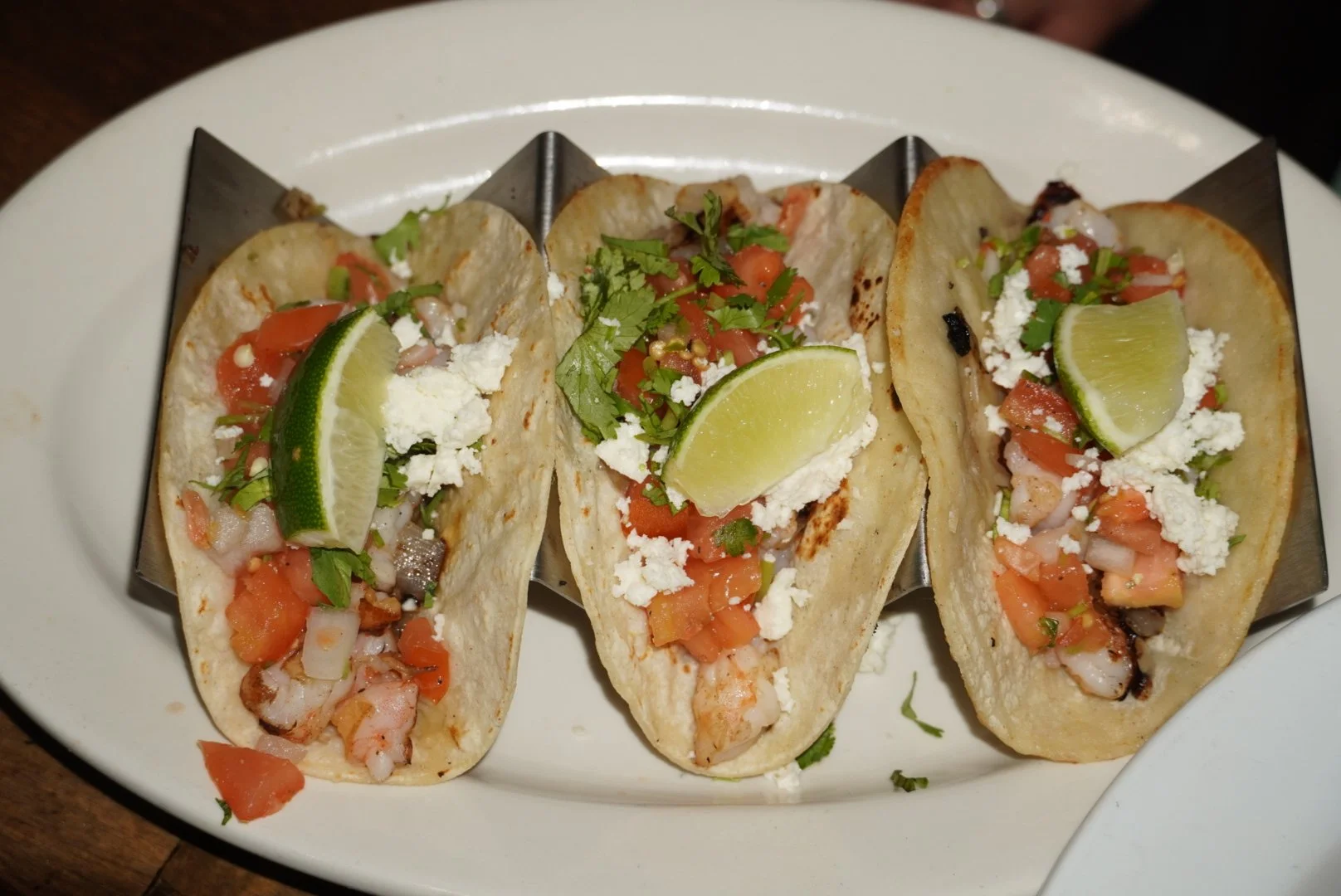 Three tacos with grilled meat, tomatoes, chopped onions, crumbled cheese, cilantro, and lime wedges on a white plate.