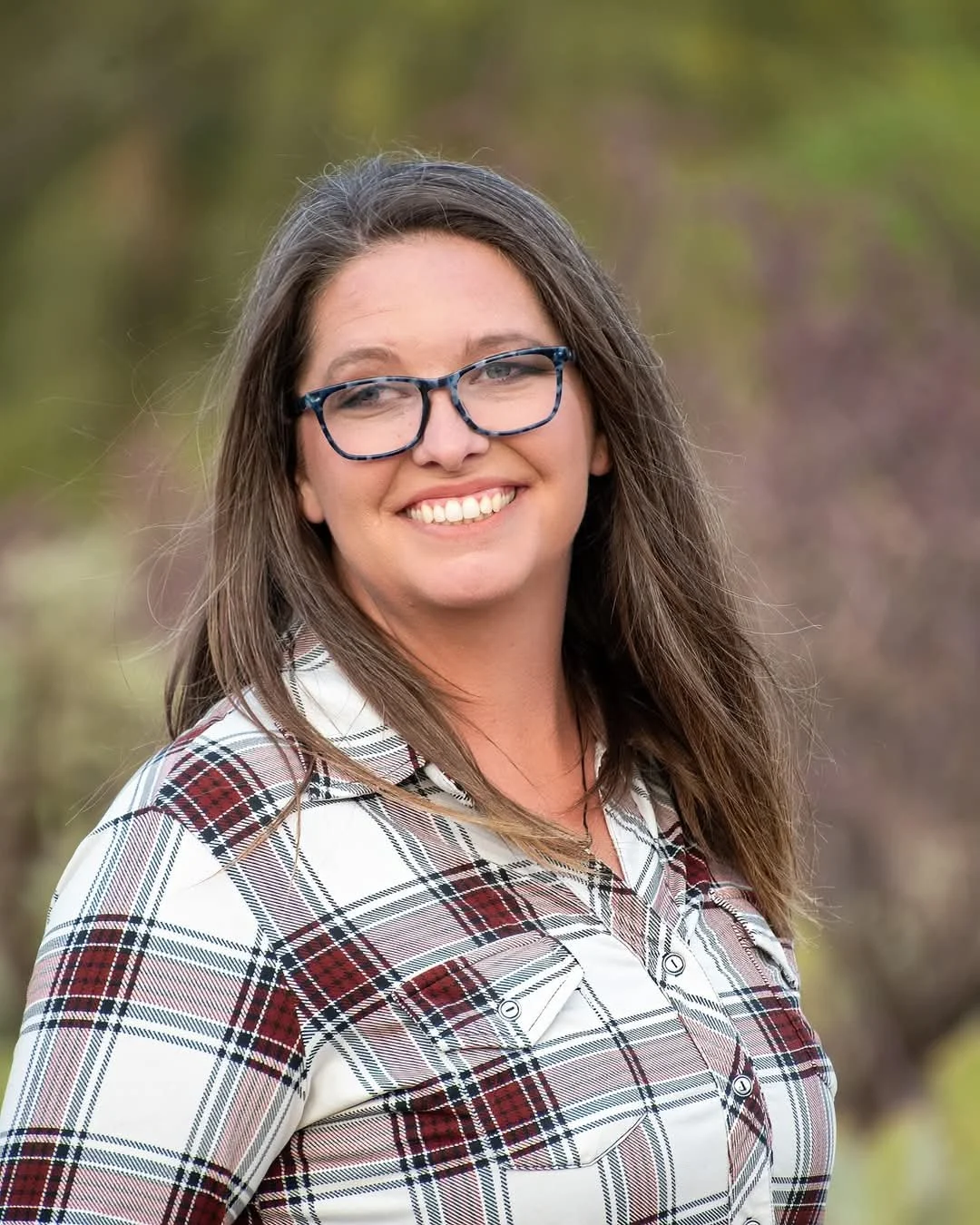 A woman with long brown hair, glasses, and a plaid shirt smiling outdoors with blurred trees in the background.
